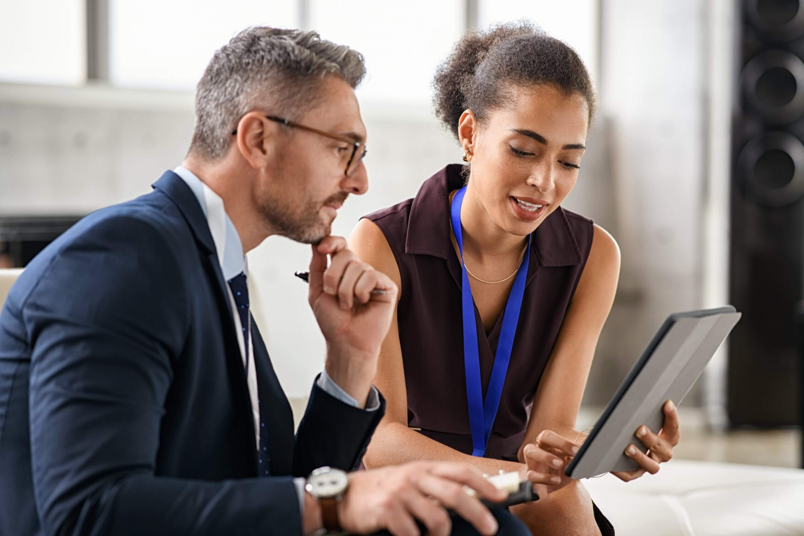 A man in a suit and a woman with a lanyard sit together, engaged in discussion. The woman holds a tablet, and they both appear focused. They are in a modern office setting with a blurred background.
