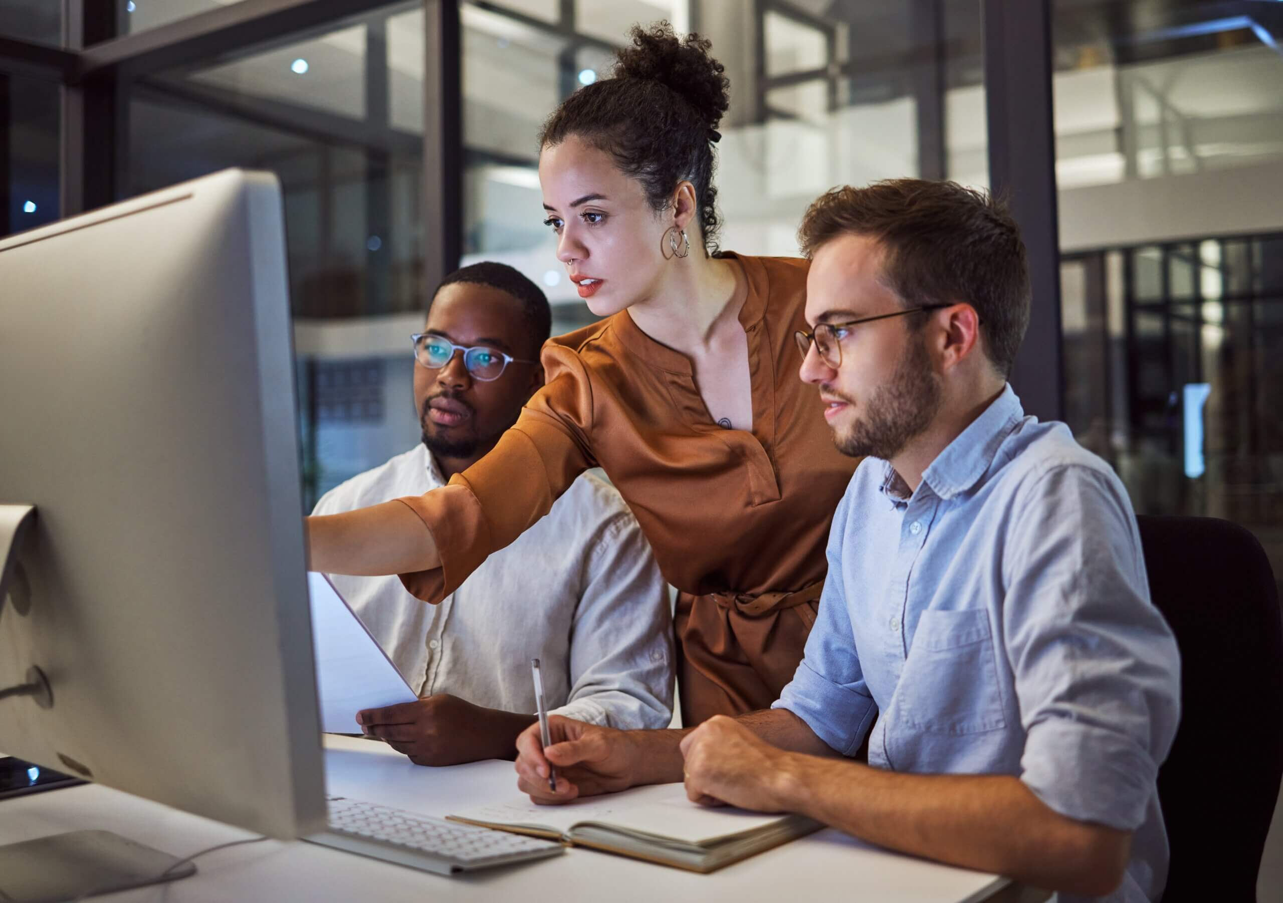 Three people working at a desk in an office. A woman stands pointing at a large computer monitor, while two men sit, one taking notes. They appear focused and engaged in a collaborative discussion.