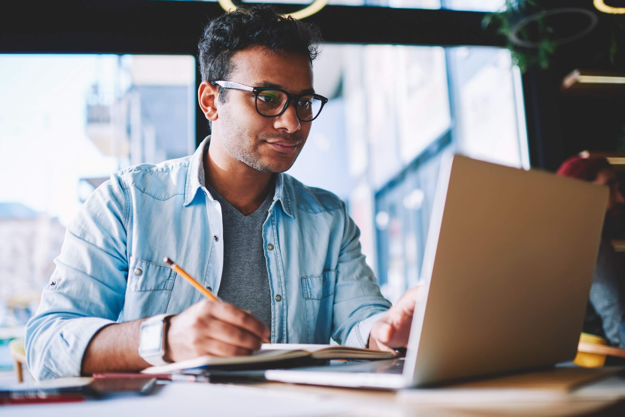 A person wearing glasses and a denim shirt is sitting at a table, working on a laptop while holding a pencil and writing in a notebook. The background shows large windows with an urban view, and the setting has a relaxed atmosphere.