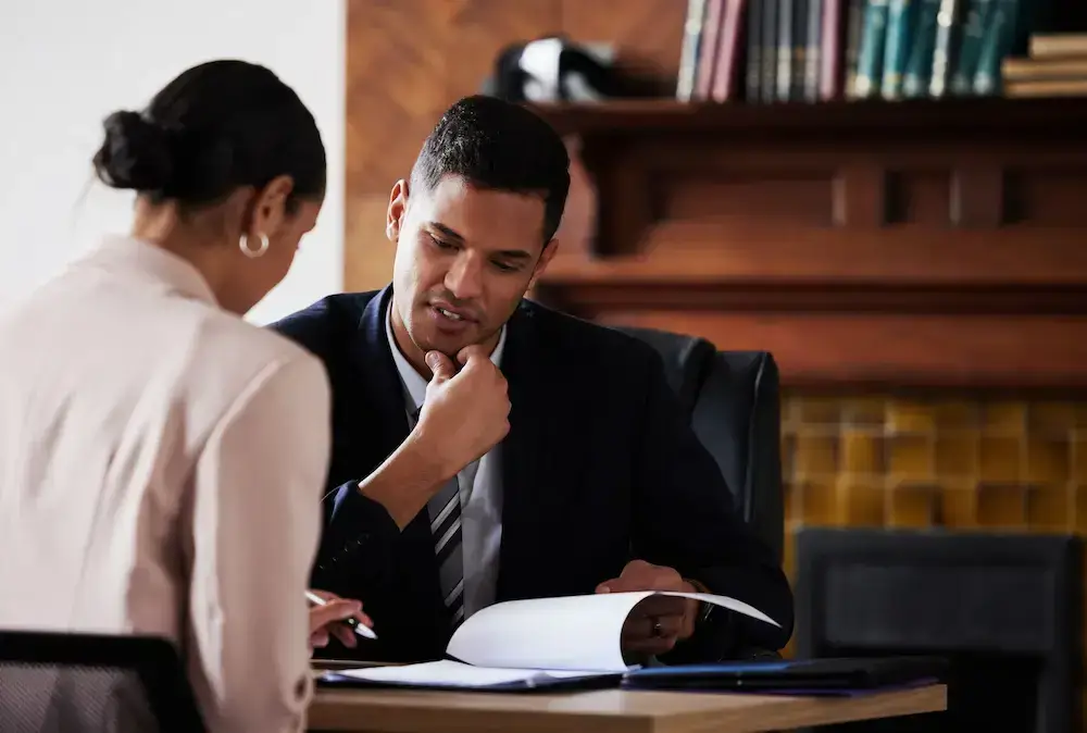 A man and a woman sit at a table with open documents. The man, wearing a suit, appears to be explaining something, while the woman listens attentively. They are in a room with a wooden mantel and a shelf with books in the background.