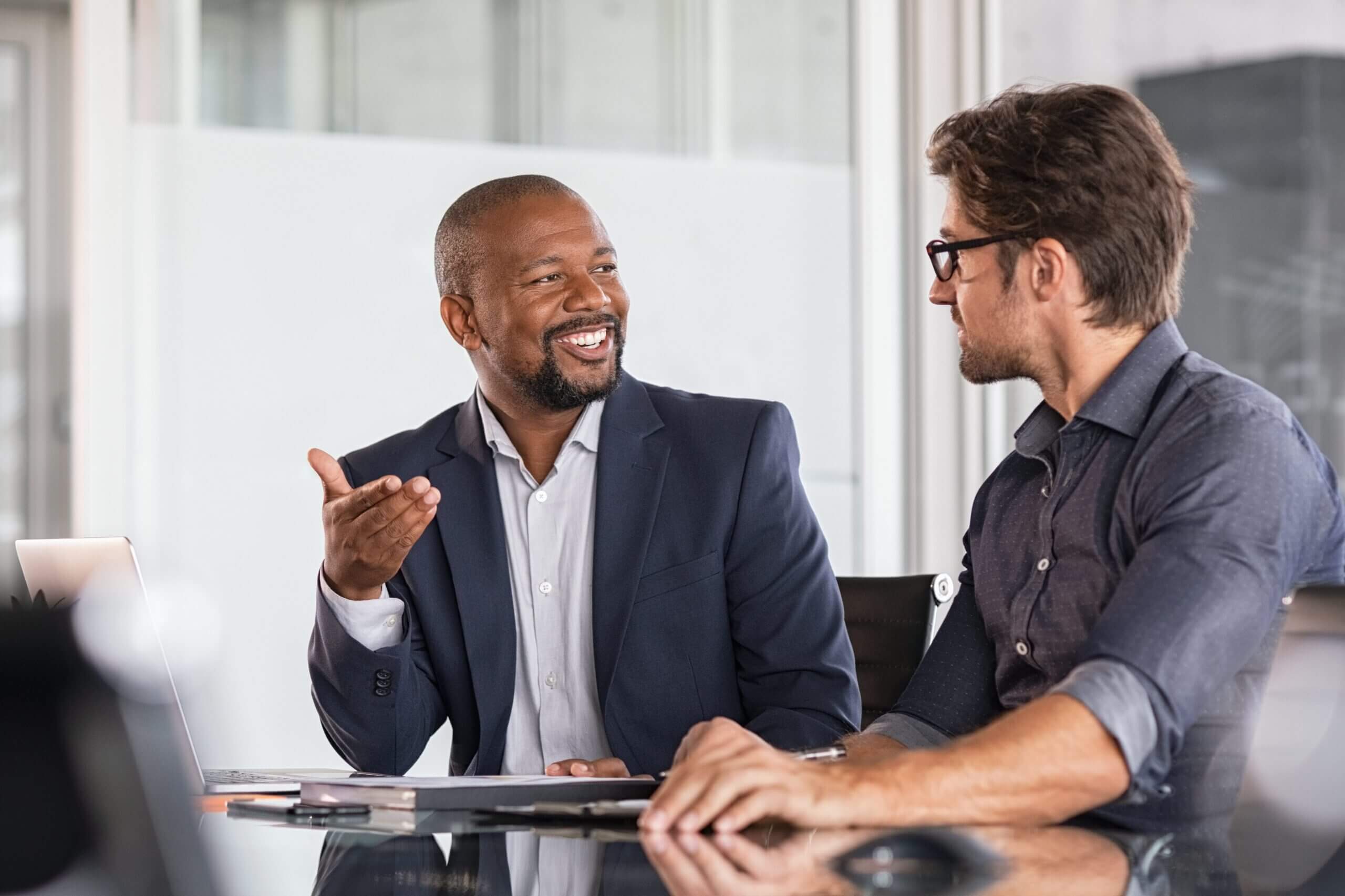 Two men sit at a conference table in an office, engaged in conversation. The man on the left is wearing a dark suit and gestures with his hand, while the man on the right, wearing glasses and a dark shirt, listens attentively.