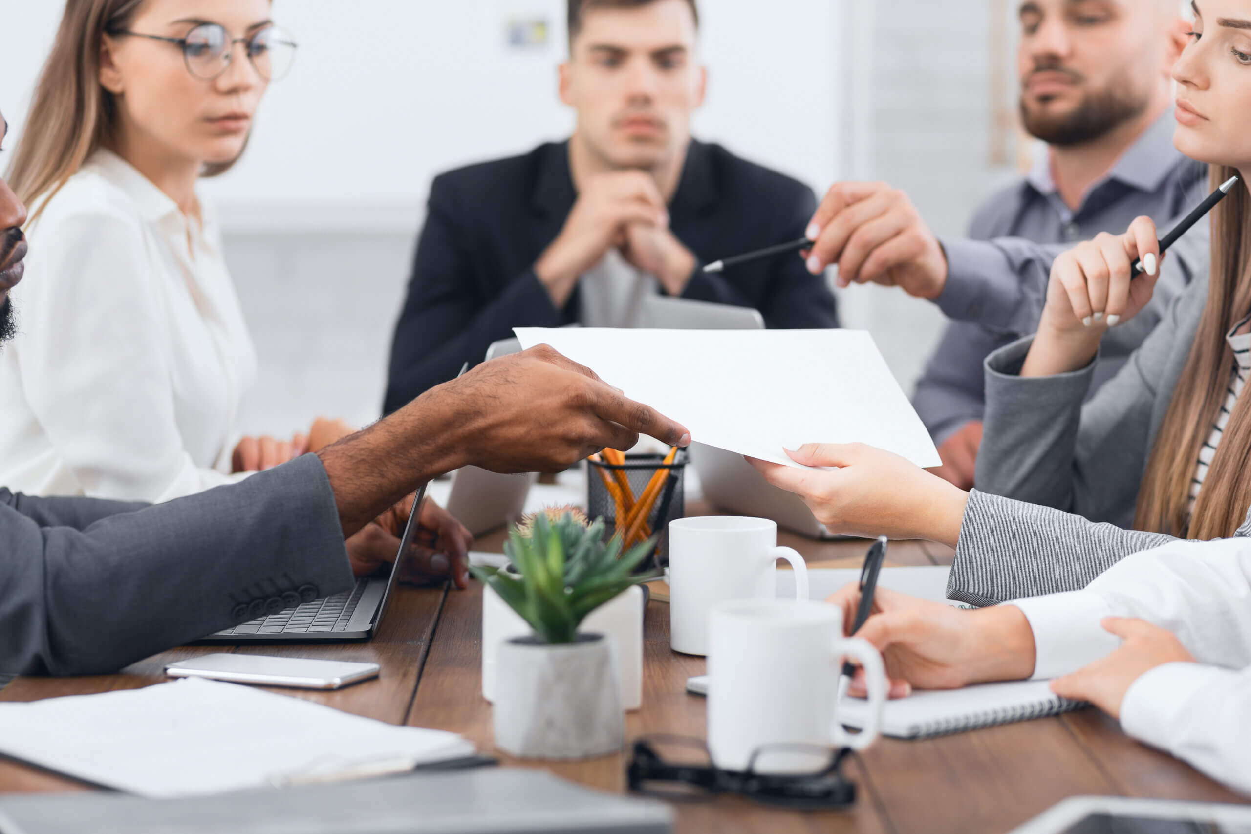 A group of people in business attire are sitting around a conference table with papers and laptops. One person is pointing to a document while others listen attentively. There are coffee mugs, notebooks, and a small plant on the table.