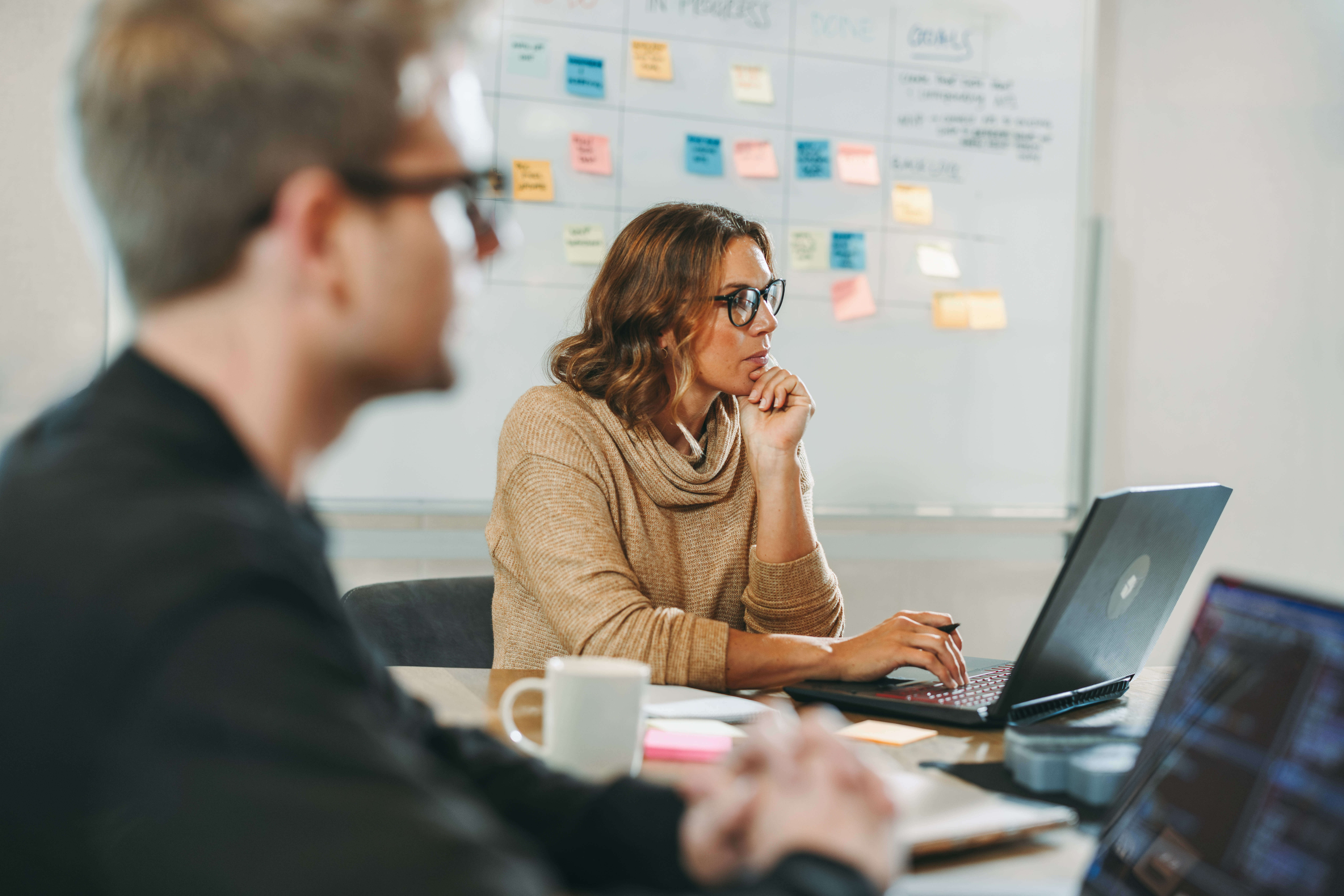 A woman wearing glasses sits at a table, focused on her laptop. A man, also wearing glasses, is blurred in the foreground. A whiteboard with colorful sticky notes is in the background. A mug and notepad are on the table.