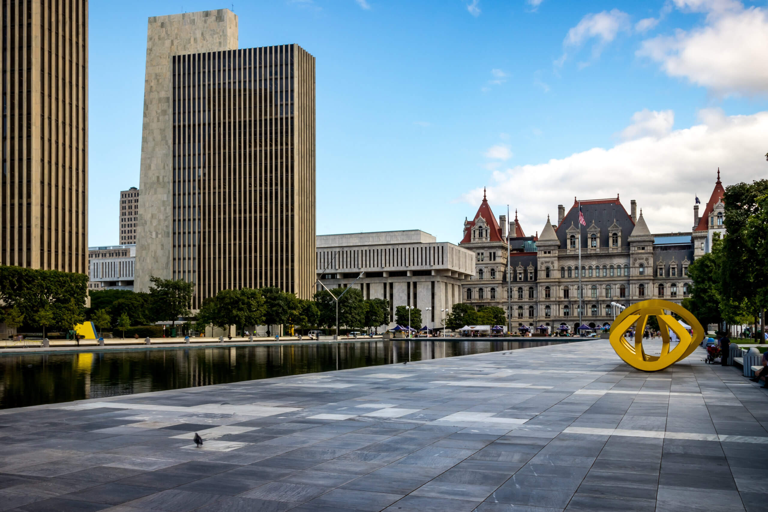 Modern urban plaza with tall rectangular buildings and a reflecting pool on the left. A large, yellow abstract sculpture is in the foreground. The background features an ornate, historic building under a partly cloudy sky.