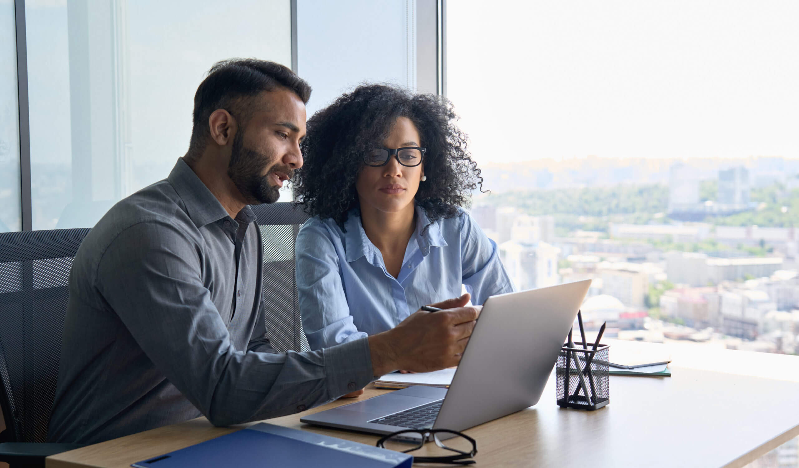 Two people sit at a desk in an office, looking at a laptop screen. The man on the left gestures towards the screen, and the woman on the right listens attentively. A pen holder and a blue folder are on the desk. A cityscape is visible outside.
