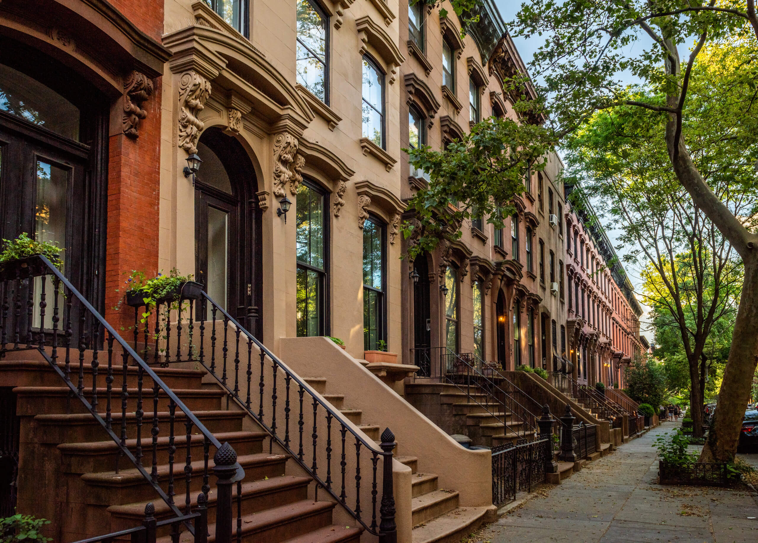 A row of historic brownstone buildings lined with trees on a residential street. Each building has an ornate facade with steps leading to the entrances. The street is calm, casting shadows from the trees as the sun sets.