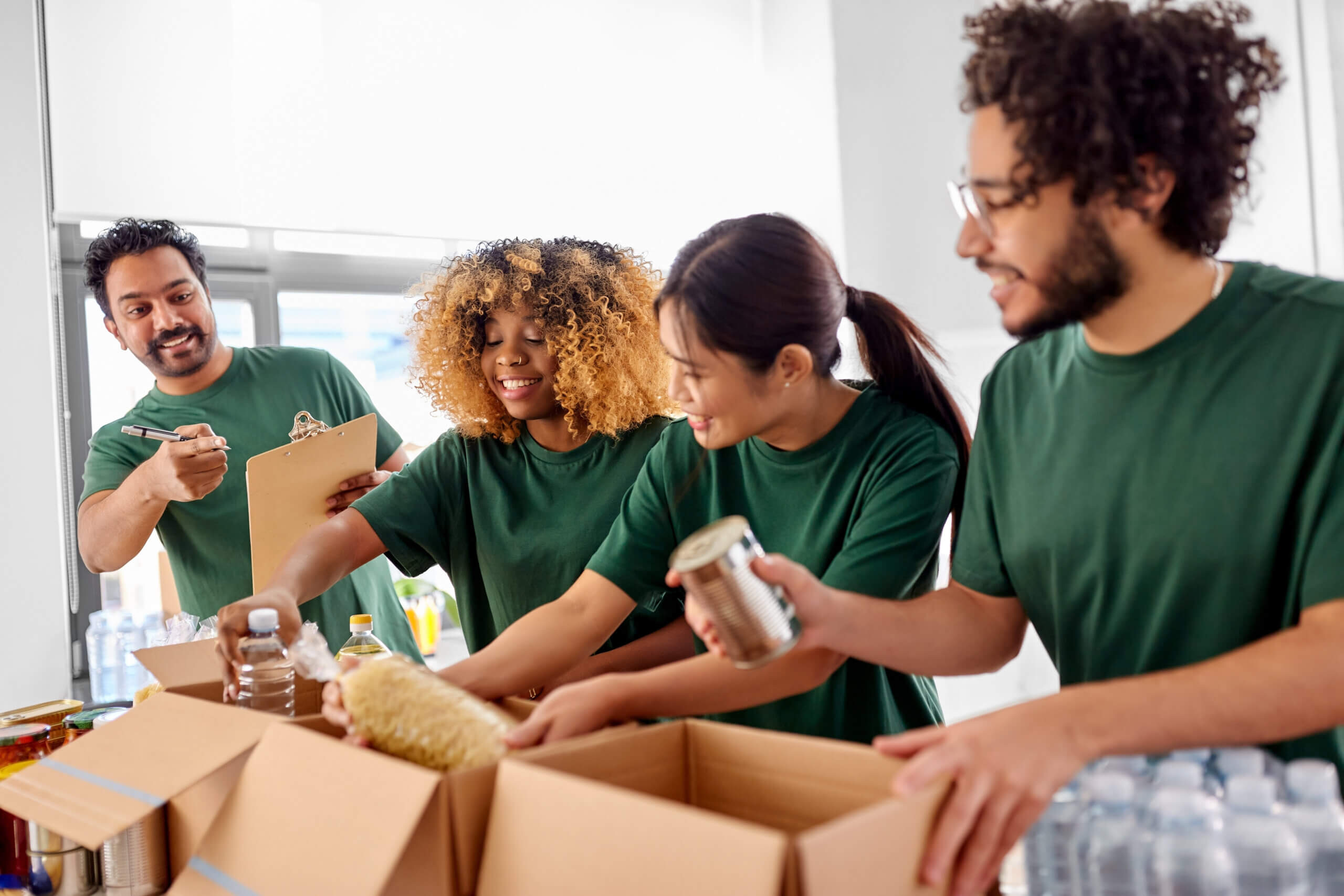 Four people wearing green shirts are packing food items into cardboard boxes. They are smiling and working together in a bright room. One person holds a clipboard, while others pack cans, bottles, and grocery items.