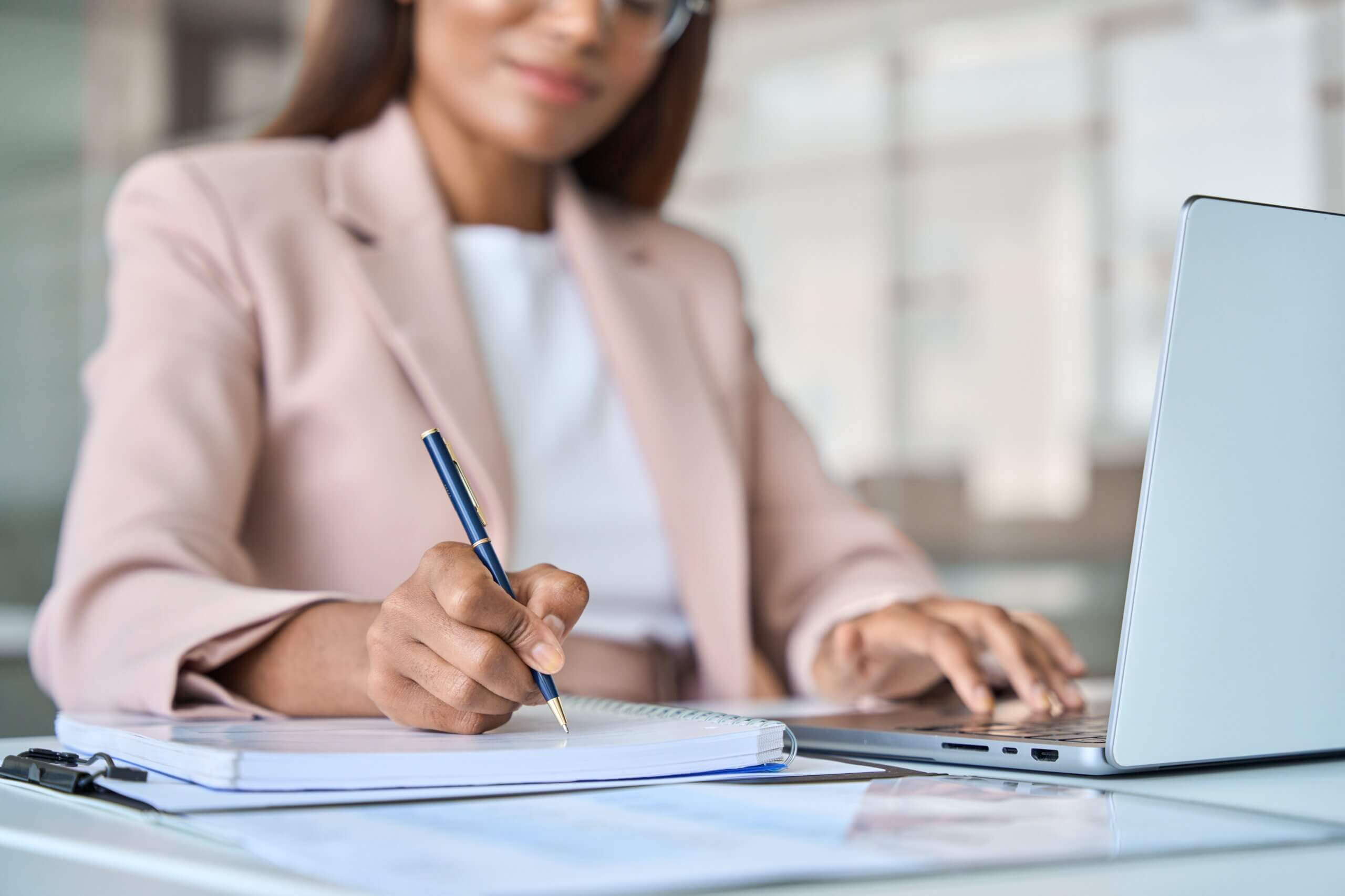 A woman in a beige blazer is sitting at a desk, writing in a notebook with a pen. She is using a laptop and appears focused on her work. The background is a blurred office setting.