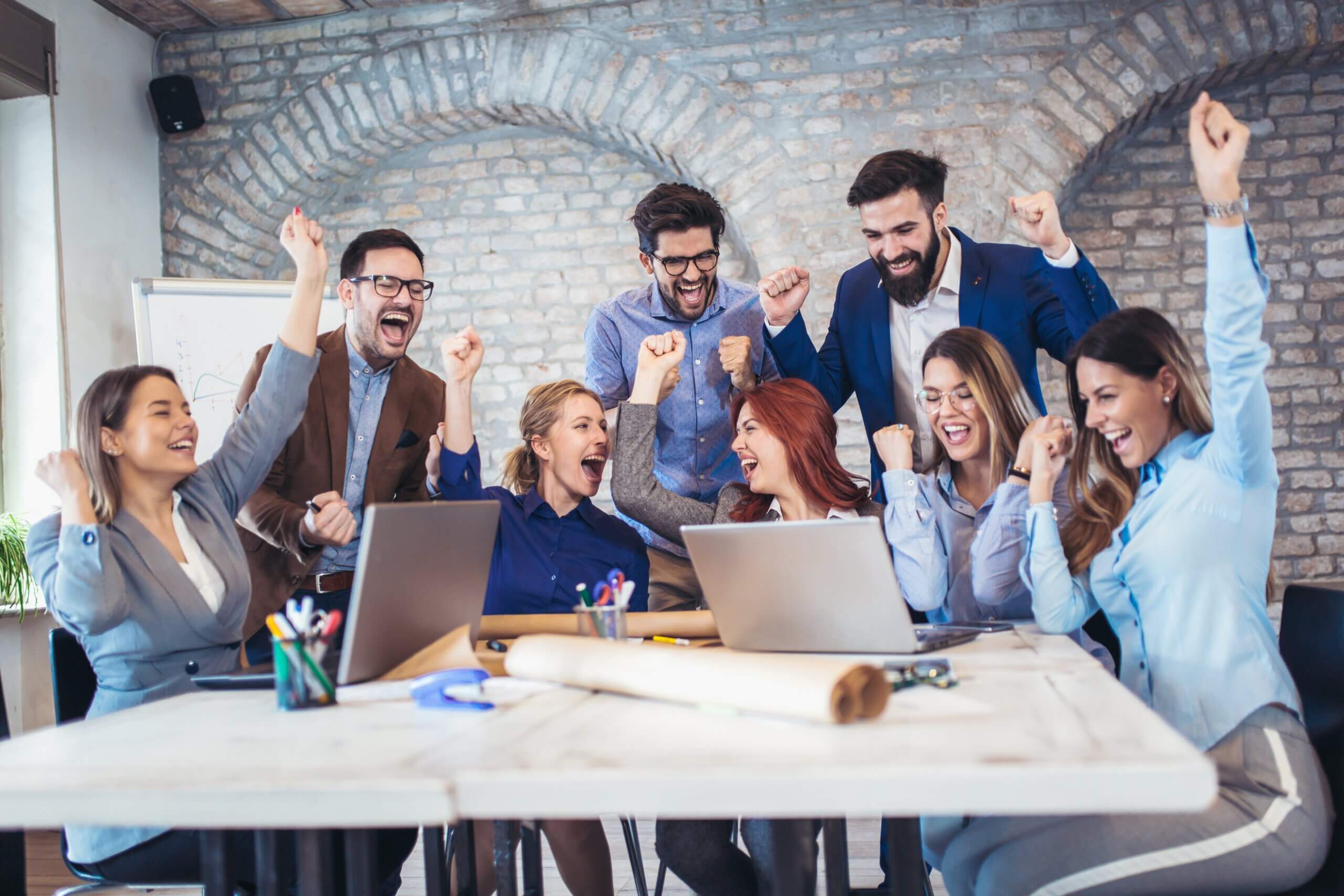 A group of people in an office celebrate around a table with laptops and papers. They are smiling, raising their arms in excitement. The setting is modern with large windows and a brick wall in the background.