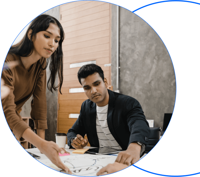 Two people collaborate at a table, examining papers and maps. One person is pointing, while the other looks on thoughtfully. A coffee cup sits on the table. They are in a modern office with a wooden door and concrete walls in the background.
