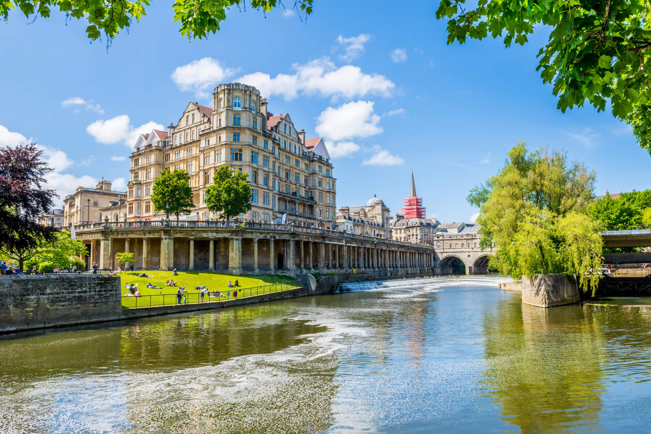 A grand, historic building with arched supports overlooks a calm river. People relax on the grassy bank. A bridge and spire in the background, surrounded by lush greenery, under a bright blue sky with scattered clouds.