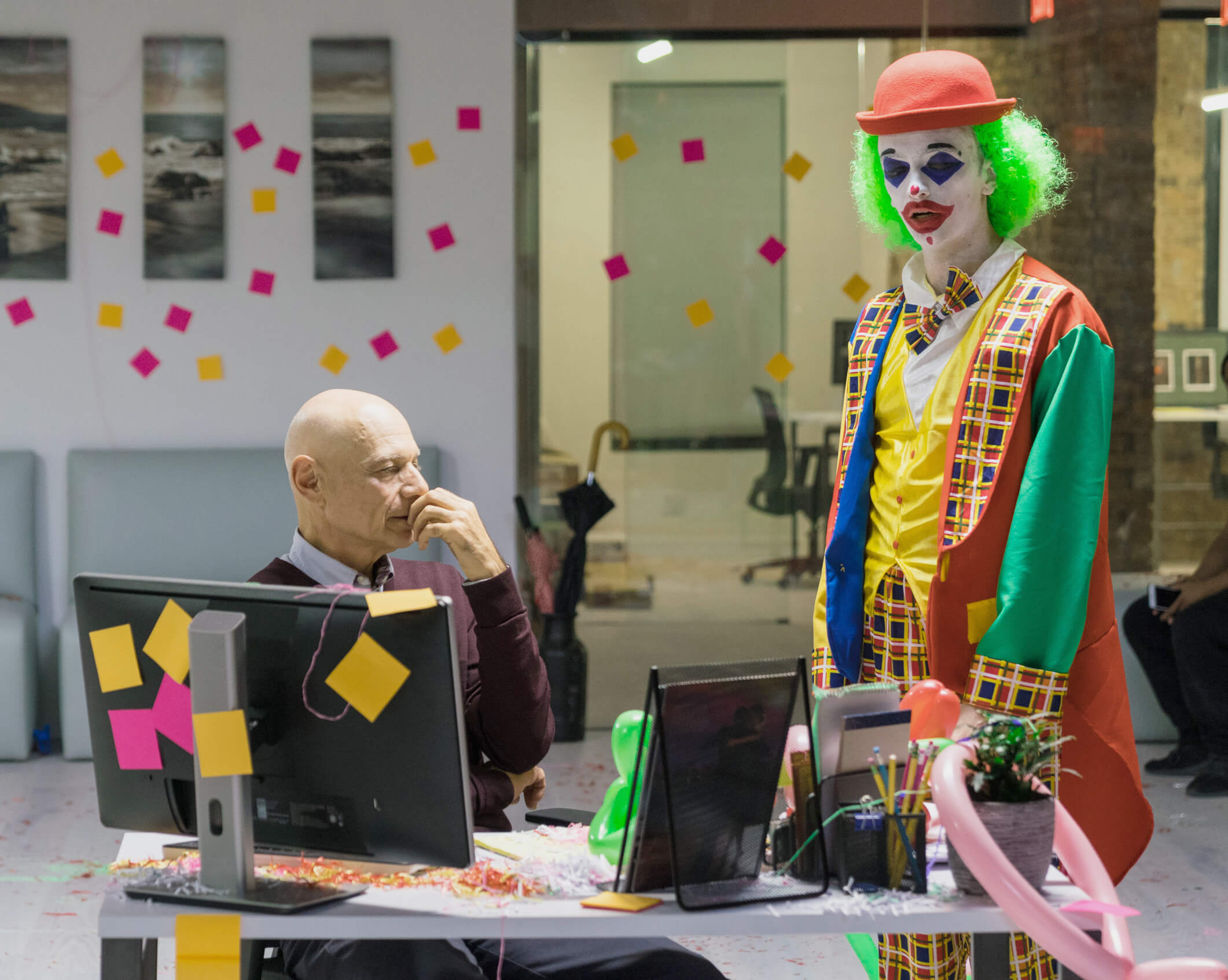 A person in a clown costume with green hair stands in an office, colorful sticky notes decorating the room. Another person sits at a desk with two monitors, looking contemplative. The atmosphere is playful and whimsical.