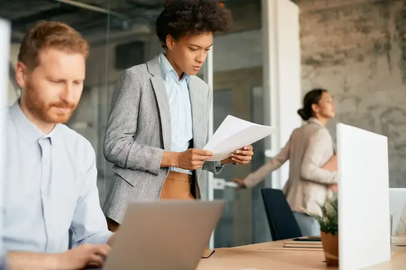 A woman in a suit reads documents in an office setting. A man in the foreground works on a laptop. In the background, another woman walks while holding a folder. The office has a modern, industrial design with a concrete wall.