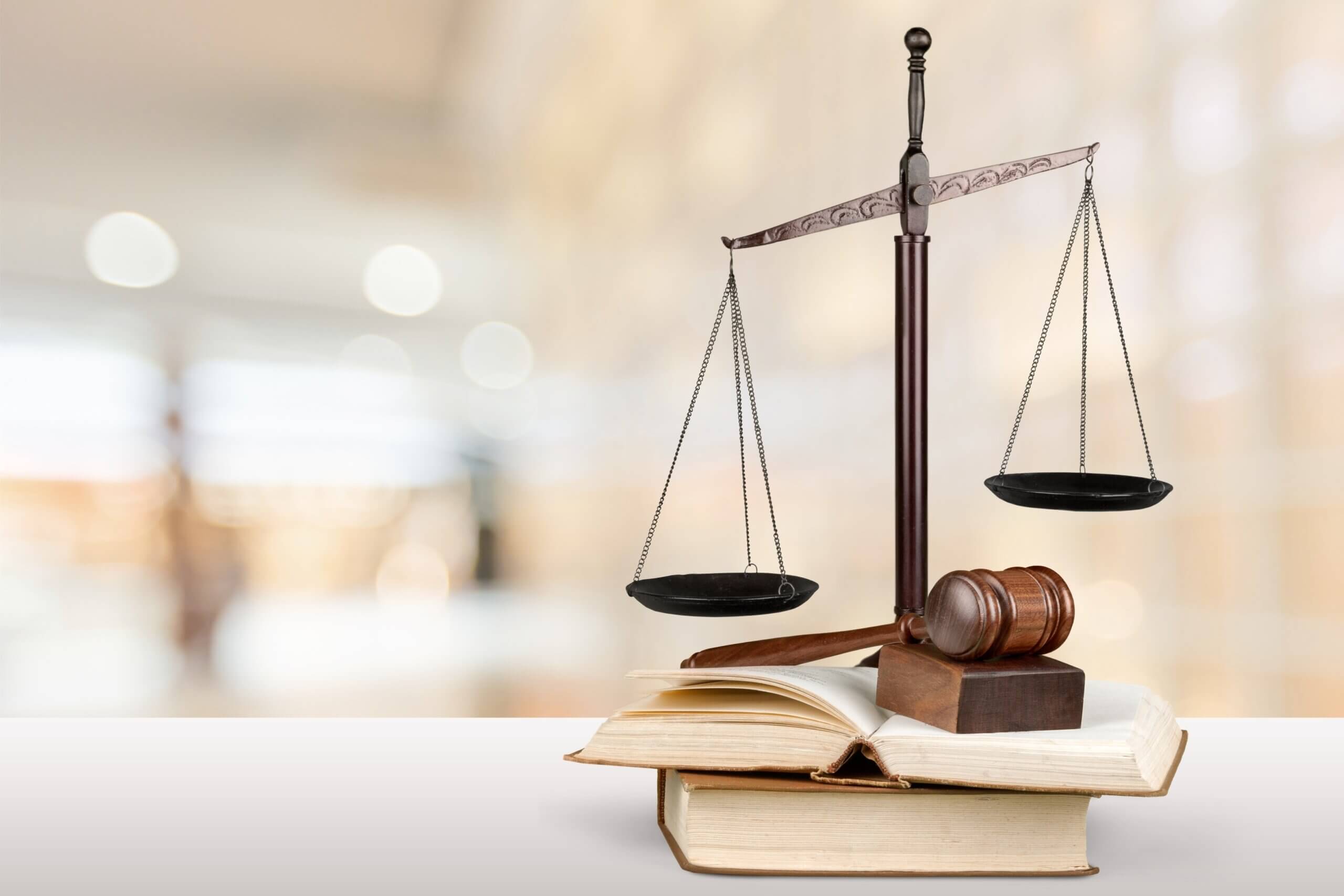 A justice scale and gavel rest on an open book. The background is softly blurred, creating a professional and scholarly atmosphere.