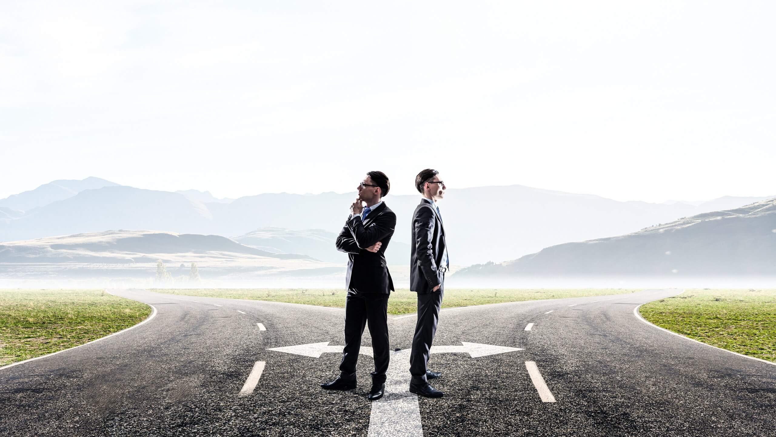 Two men in suits stand back-to-back at a crossroads on an open road. Each faces a different direction, suggesting decision-making. The background shows misty hills and a clear sky.