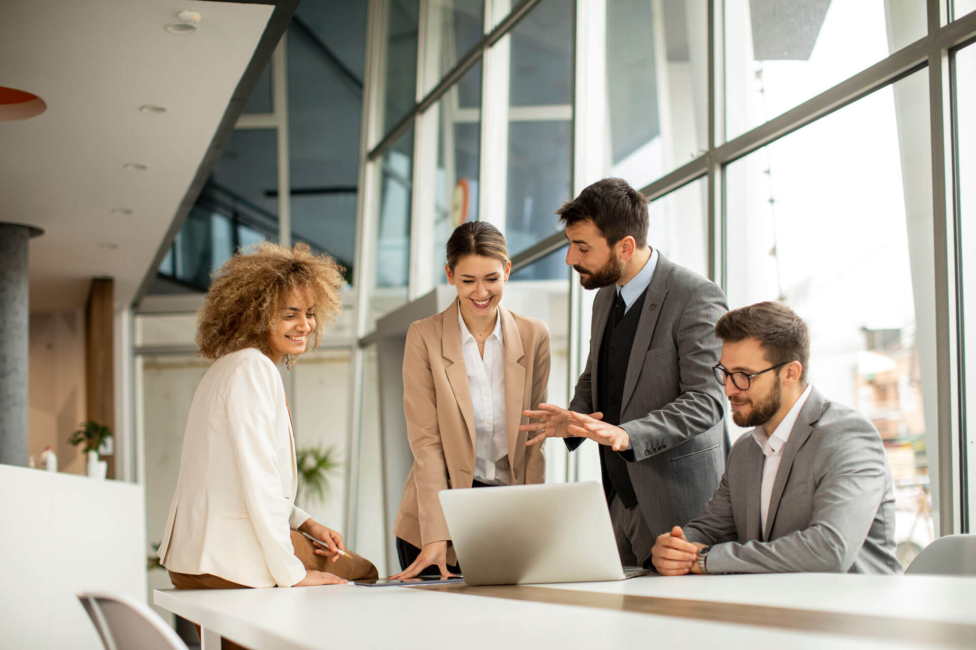 Four people in business attire are engaged in a discussion around a laptop in a modern office with large windows. One person is explaining something while others listen attentively, representing Cogency Global’s hands-on company formation services.