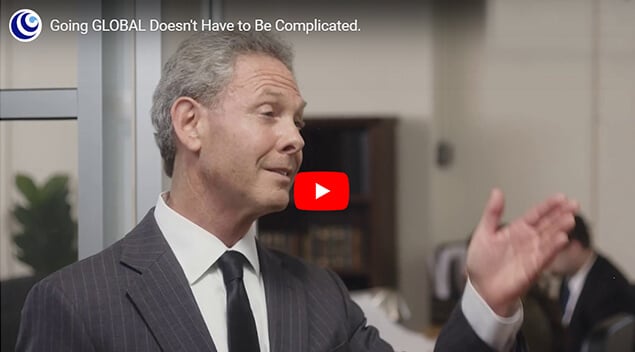 A man in a suit gestures while speaking in an office setting. Another person is visible in the background, seated at a desk. Text at the top of the image reads, "Going GLOBAL Doesn't Have to Be Complicated.