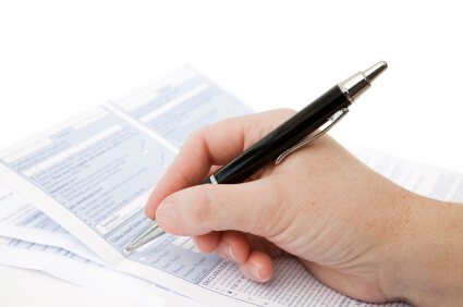 A hand holds a pen over a form with several documents layered beneath it on a white surface. The scene suggests someone is in the process of filling out paperwork.