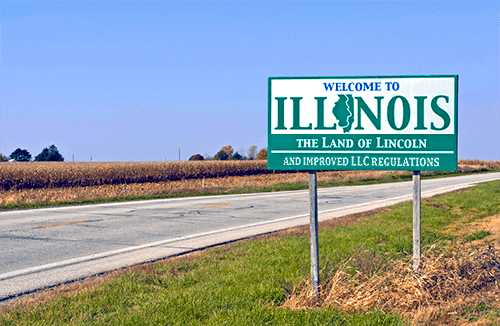 A roadside sign reads "Welcome to Illinois, The Land of Lincoln and Improved LLC Regulations." It stands by an empty highway with a cornfield and a clear blue sky in the background.