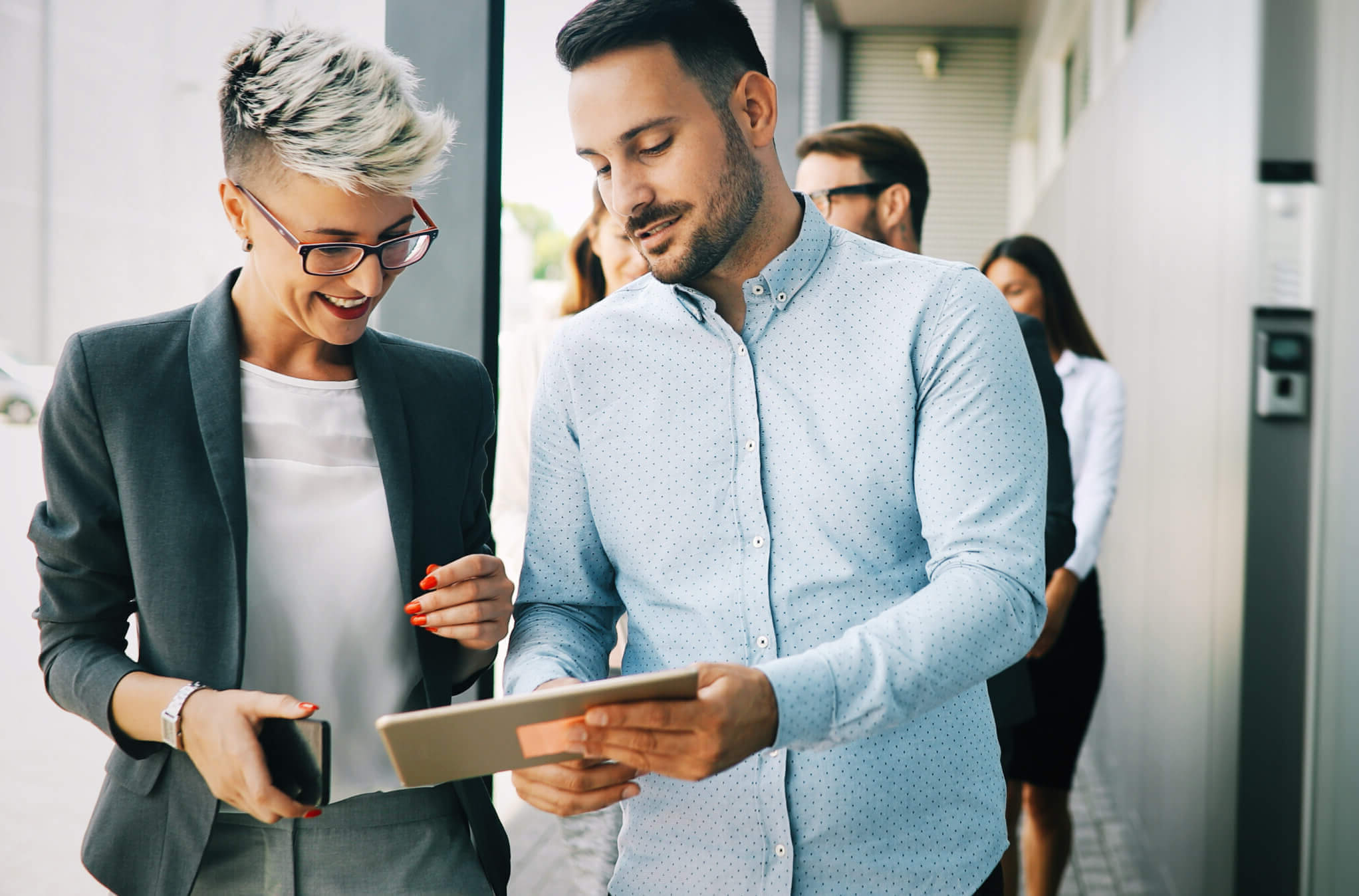 A man and woman in business attire are standing outside a building, smiling and looking at a tablet. The woman holds a smartphone. Two other people are in the background.