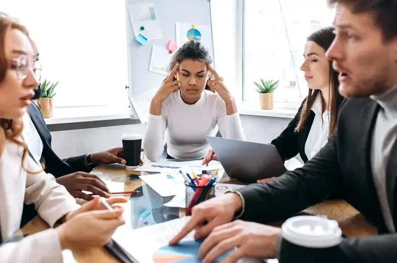 A group of five people sit around a table in a meeting room. One person looks stressed with fingers on temples. Others engage in discussion. Papers, charts, and a laptop are on the table. A window and plants are in the background.