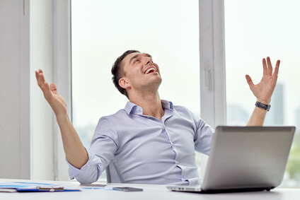 A man in a light blue shirt sits at a desk with a laptop, looking upward with a joyful expression and raised arms. He is inside a bright room with large windows, and a few papers are spread on the desk.