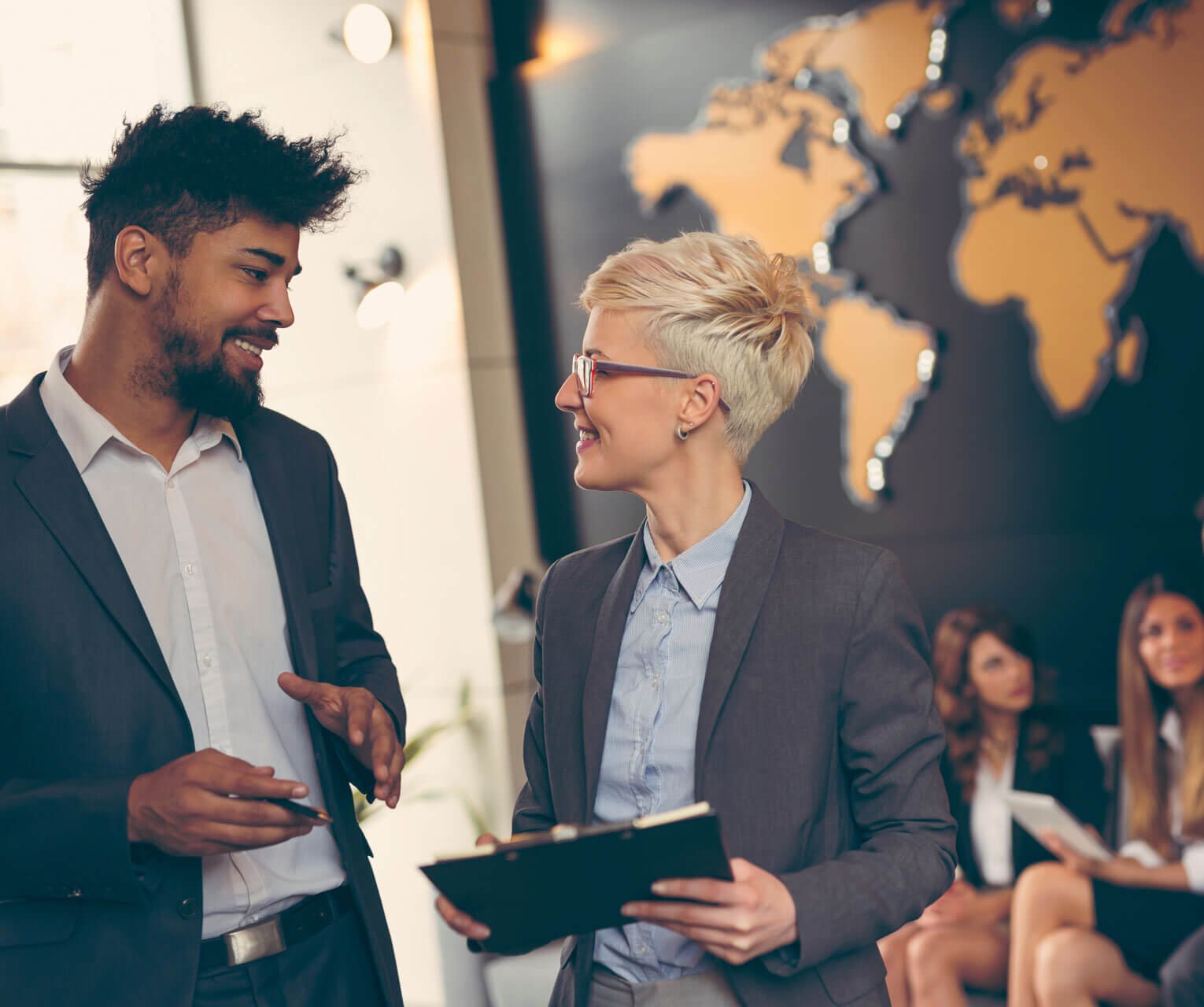 A man and a woman in business attire are talking and smiling in an office setting. The woman holds a clipboard, representing their focus on nonprofit compliance. A world map is displayed on the wall behind them.