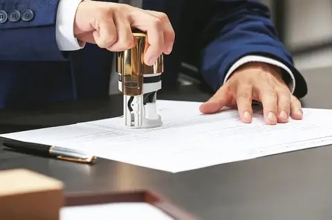 A person in a blue suit uses a round stamp on a document placed on a desk. A pen lies on the table next to the paper. The setting appears to be a professional or office environment.