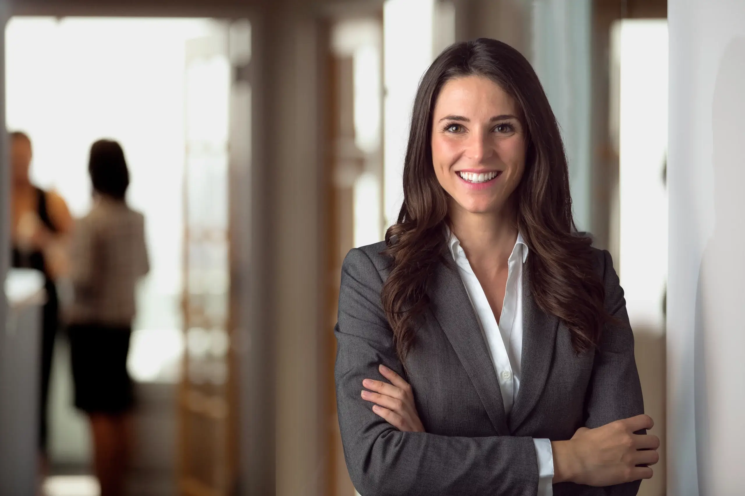 A woman in a gray blazer and white blouse stands smiling with arms crossed in an office hallway. She has long dark hair. In the blurred background, two people are conversing near a doorway.