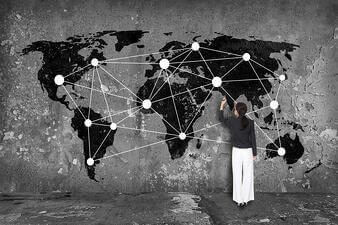 A person in a dark blazer and white pants stands facing a large wall mural of a world map. They are adding lines connecting white circles on the map, illustrating a network across various continents.