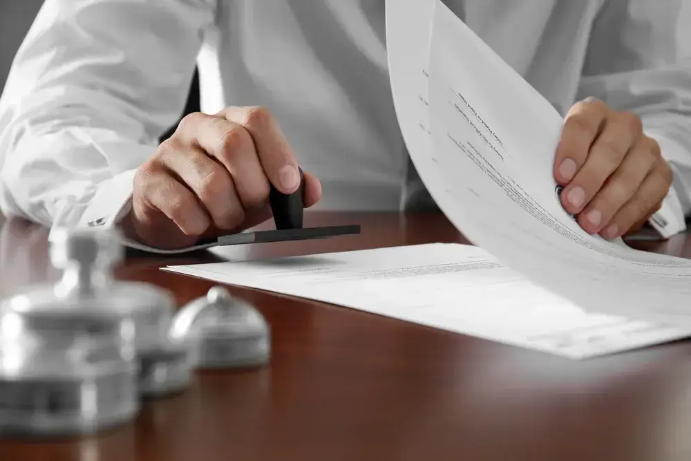 A person in a white shirt is stamping documents on a wooden desk. Several stamps are visible nearby. The image focuses on the hands and paperwork, conveying an official or administrative action.