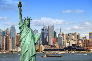 The Statue of Liberty stands prominently with New York City skyscrapers in the background under a clear blue sky. A boat is visible in the water in front of the cityscape.