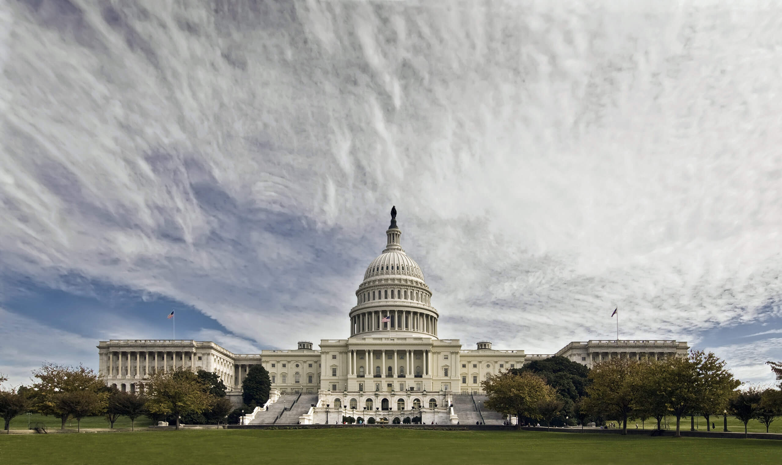 The image shows the United States Capitol building under a dramatic, cloudy sky. The neoclassical structure features a large central dome, flanked by columns and wings. Trees and a grassy area are in the foreground.