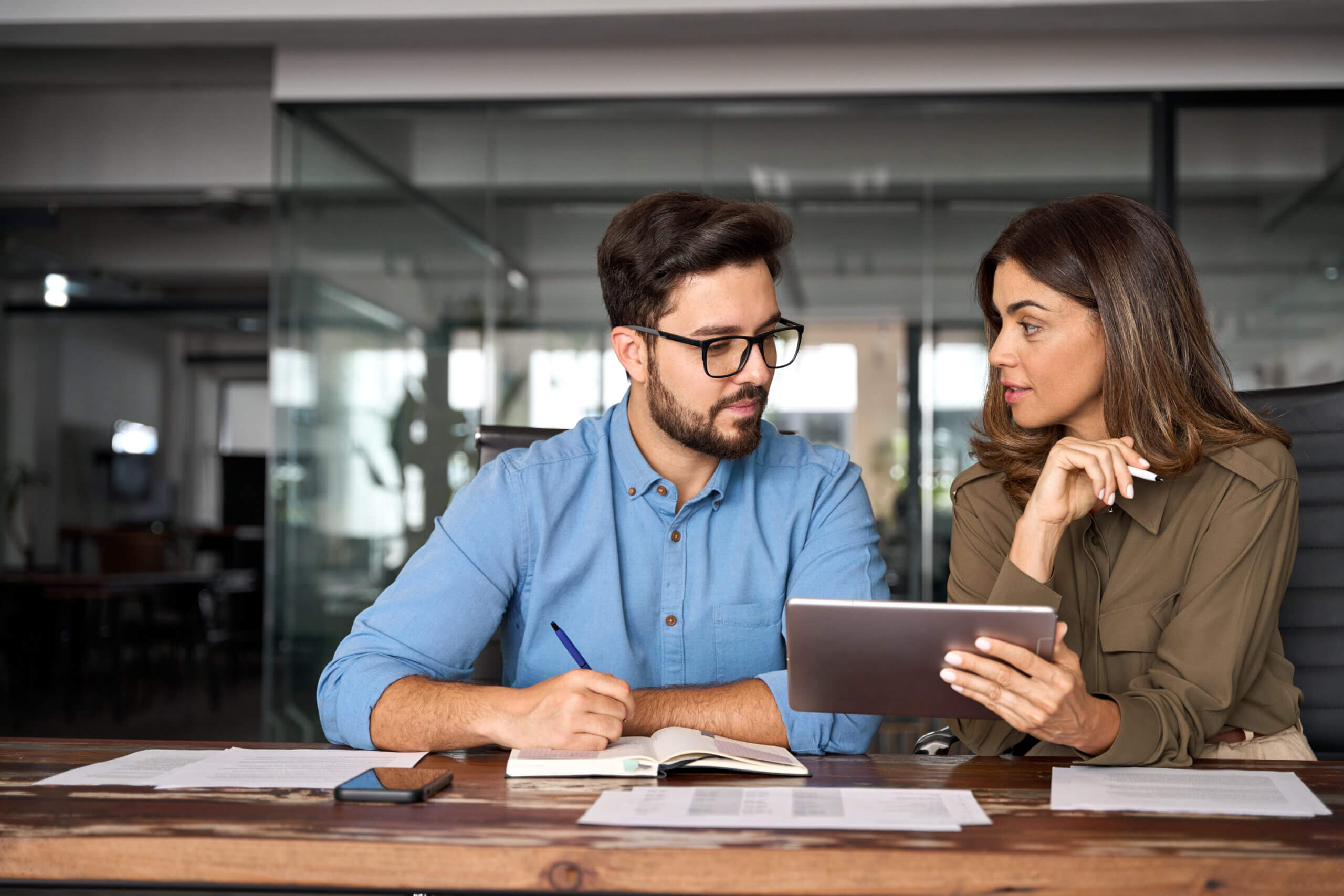 A man and woman are sitting at a desk in an office. The man in a blue shirt is taking notes in a notebook. The woman in a green shirt is holding a tablet, showing something to the man. Papers and a phone are on the desk.