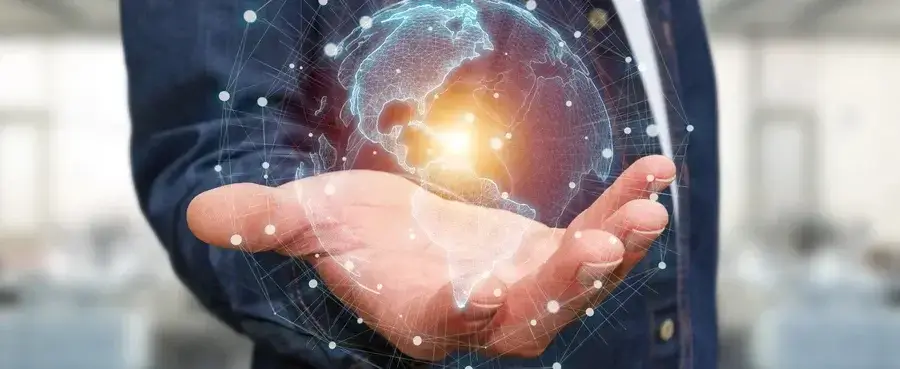 A person in a blue jacket holds out their hand with a glowing holographic image of the Earth above it. The hologram displays a network-like pattern with bright light at the center. The background is blurred and indistinct.