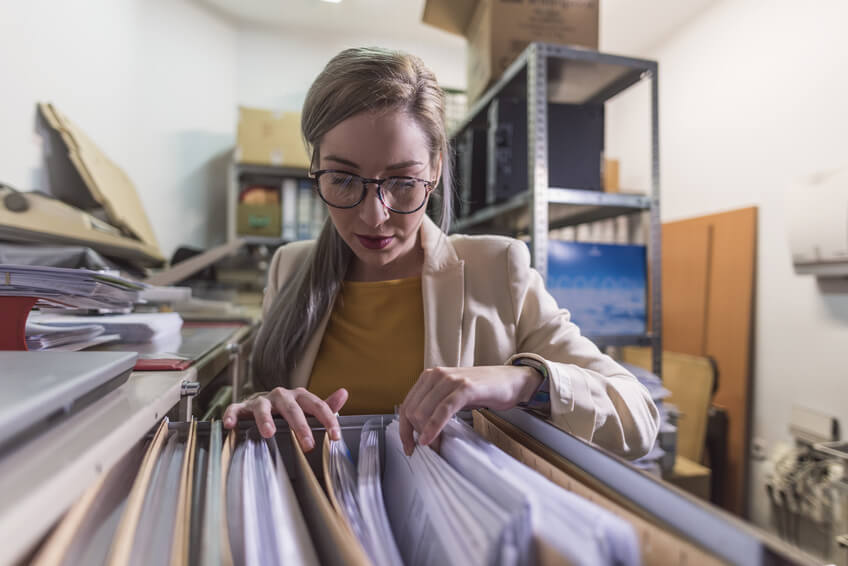A person wearing glasses and a beige blazer is looking through files in a cabinet in a cluttered office storage room. Shelves with boxes and documents are visible in the background.