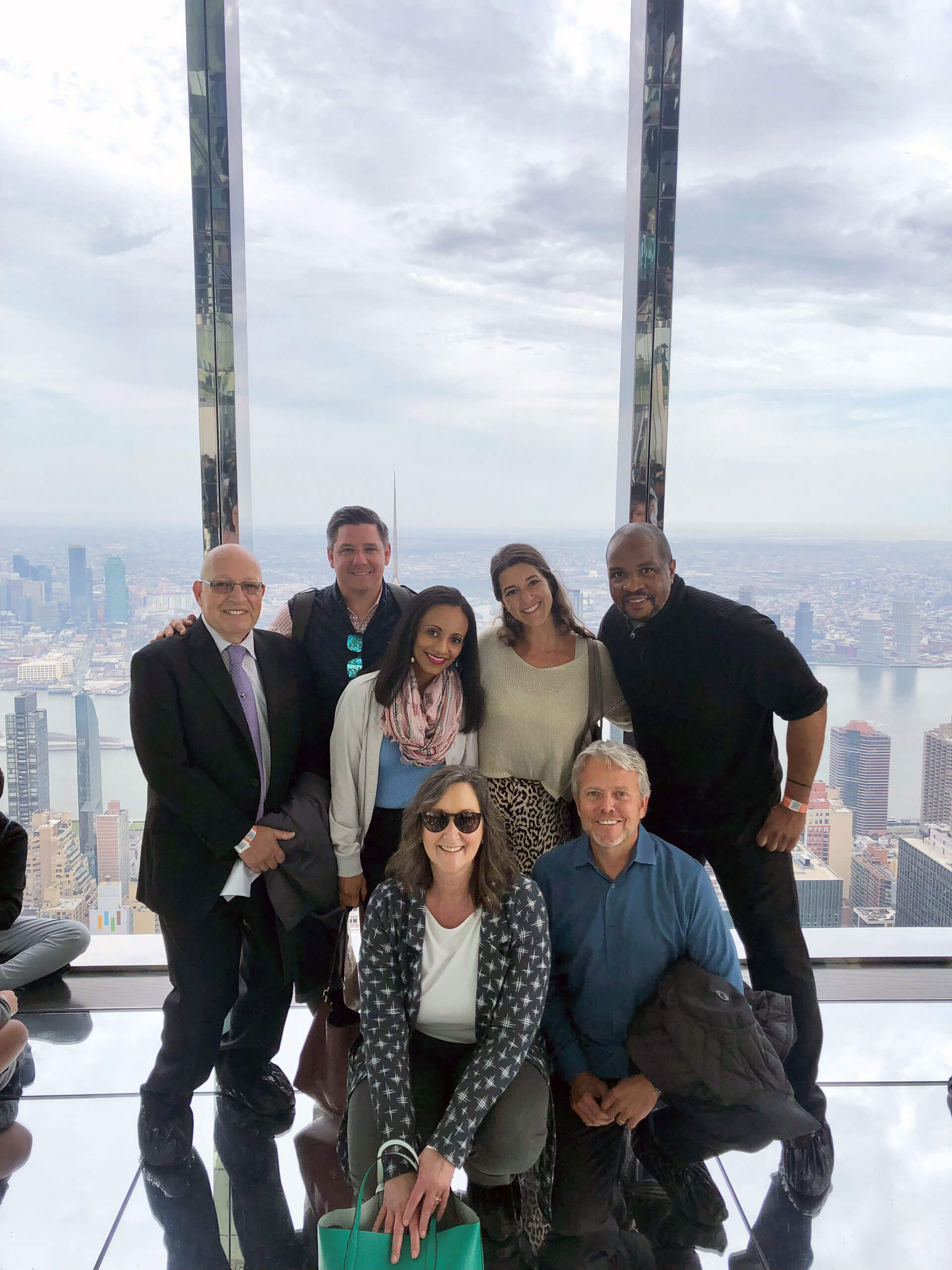A group of seven people posing and smiling inside a glass building with a cityscape view in the background. They are wearing a mix of casual and business attire. The skyline and cloudy sky can be seen through large windows.