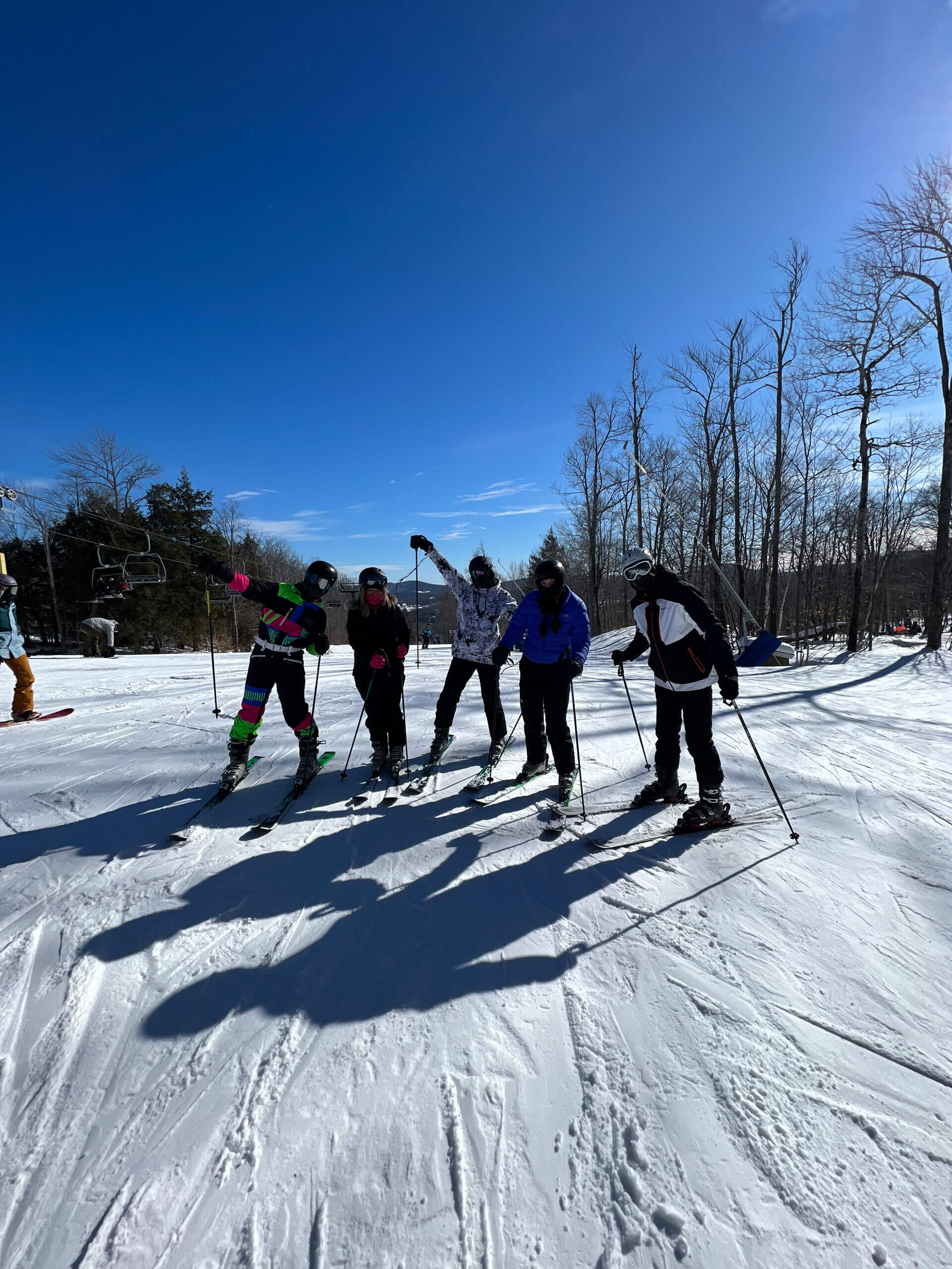 Five people in ski gear standing on a snowy slope, under a clear blue sky. They're posing with their ski poles, with leafless trees and a ski lift in the background, casting long shadows on the snow.