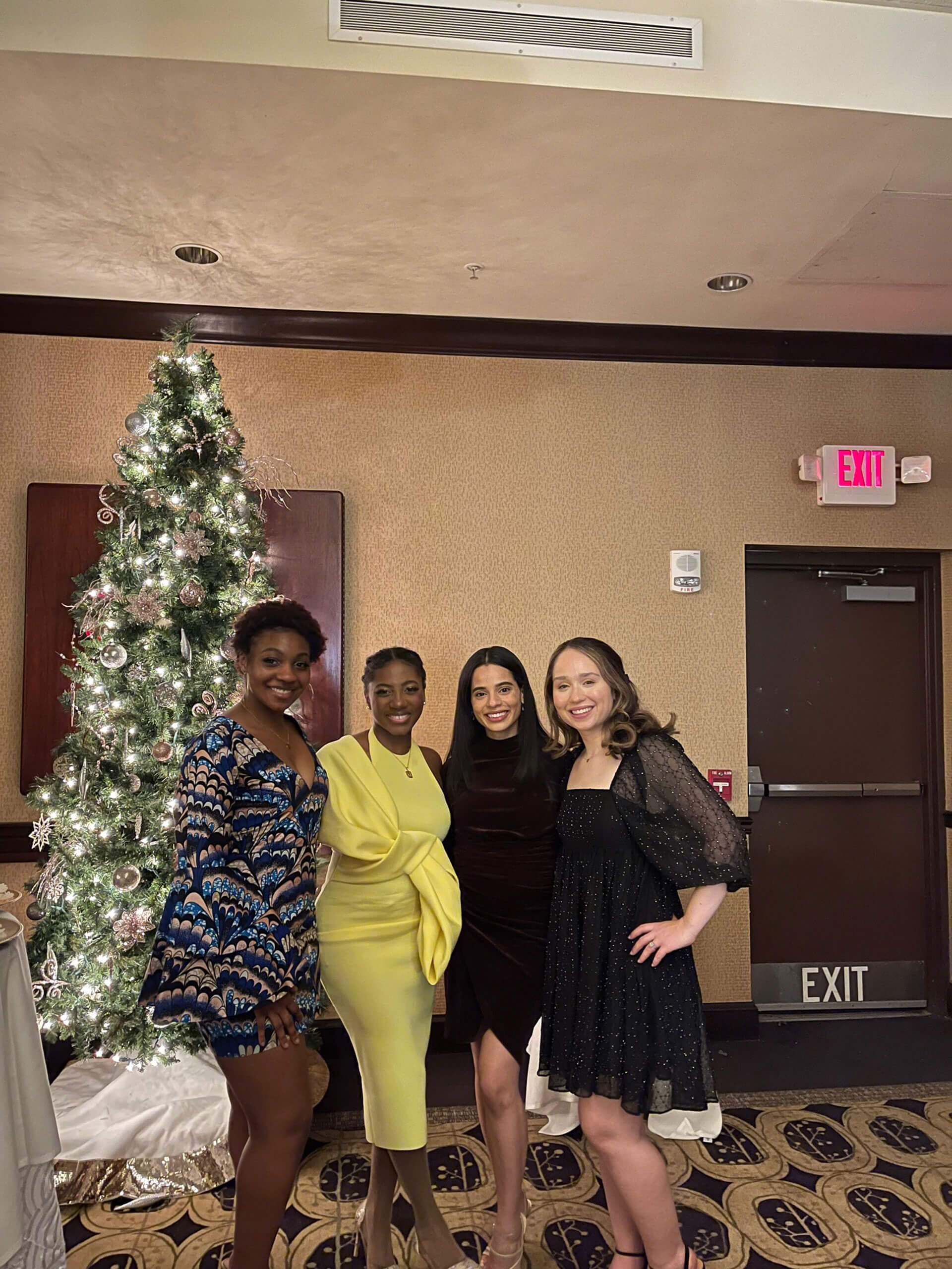 Four women are posing together in a festive setting. A decorated Christmas tree stands behind them, and they're dressed in elegant party attire. The room has warm lighting and an exit sign visible in the background.