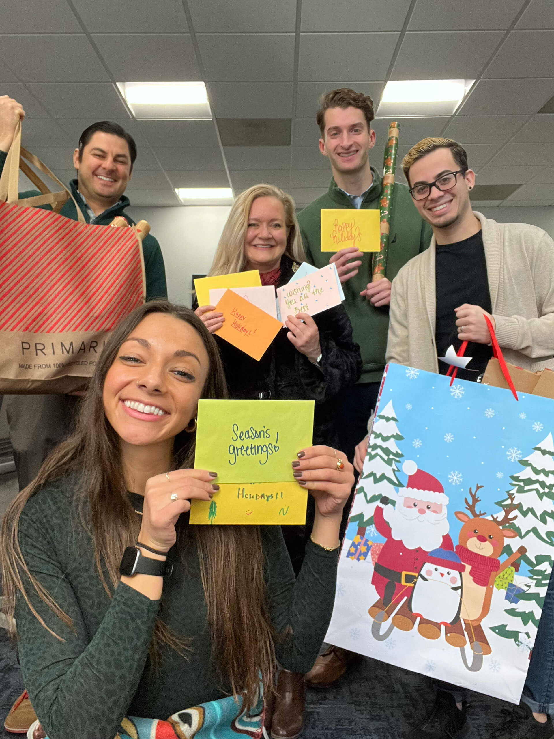 A group of five people smiling and holding holiday cards and gift bags. One person holds a card with "Season's Greetings" written on it. Another person holds a large gift bag with a Santa and reindeer design. They appear to be celebrating the holidays.