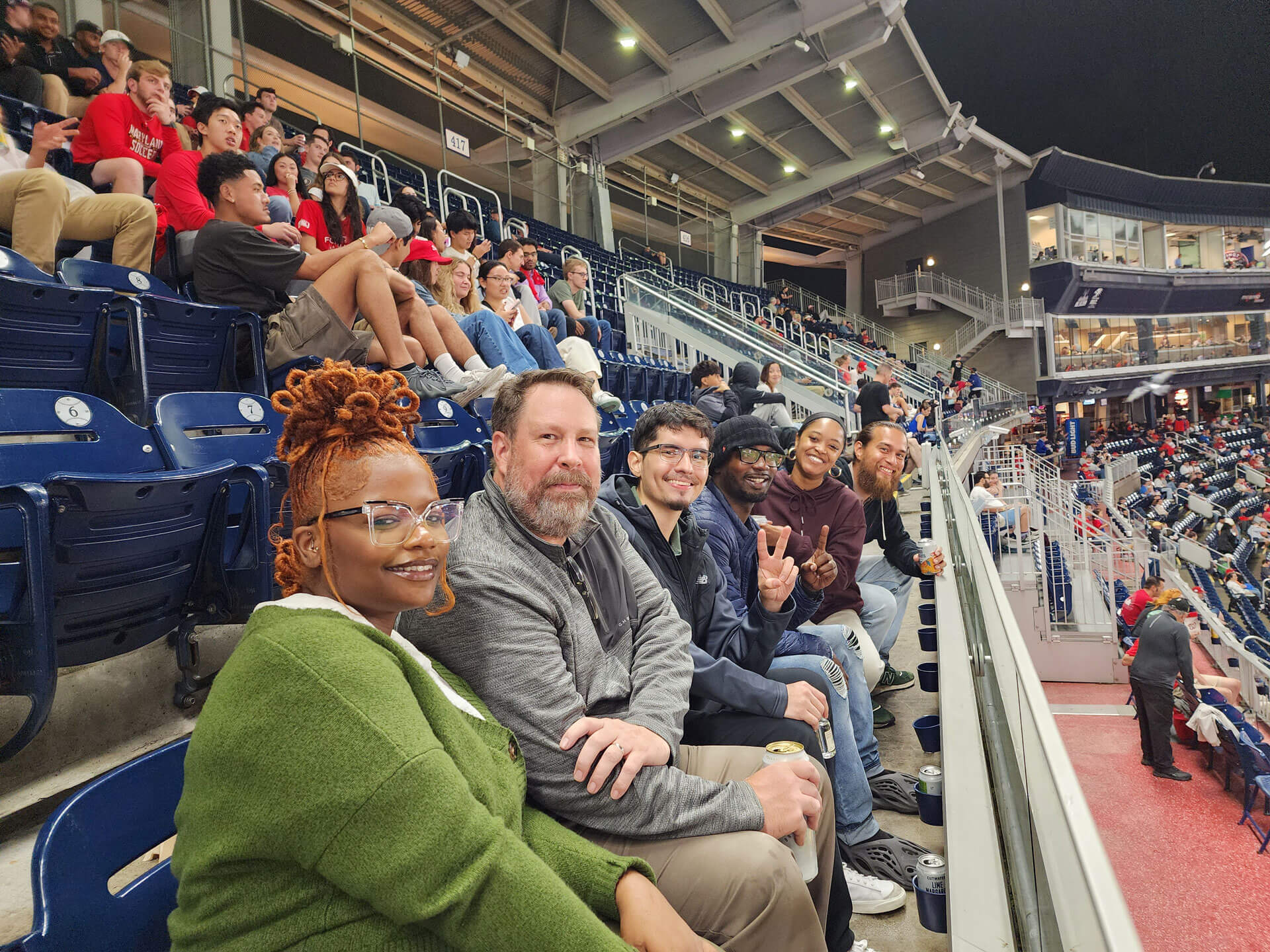 A group of six people, seated in a row, smiles for the camera at a sports stadium. Behind them, more spectators sit in the stands, watching the event. The scene captures a lively atmosphere with bright stadium lights overhead.