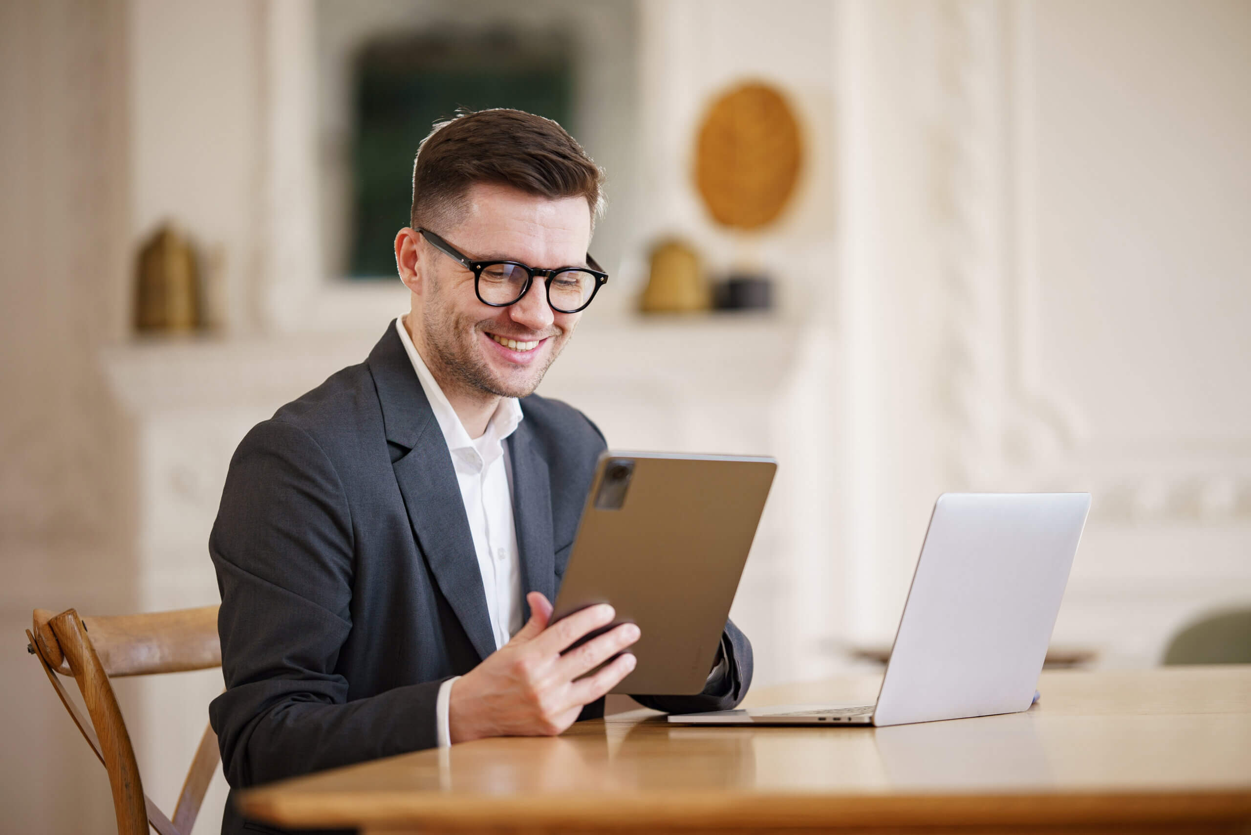 A man in a suit and glasses is smiling while holding a tablet. He is seated at a wooden table with a laptop in front of him. The background is a softly decorated room with shelves and framed elements.