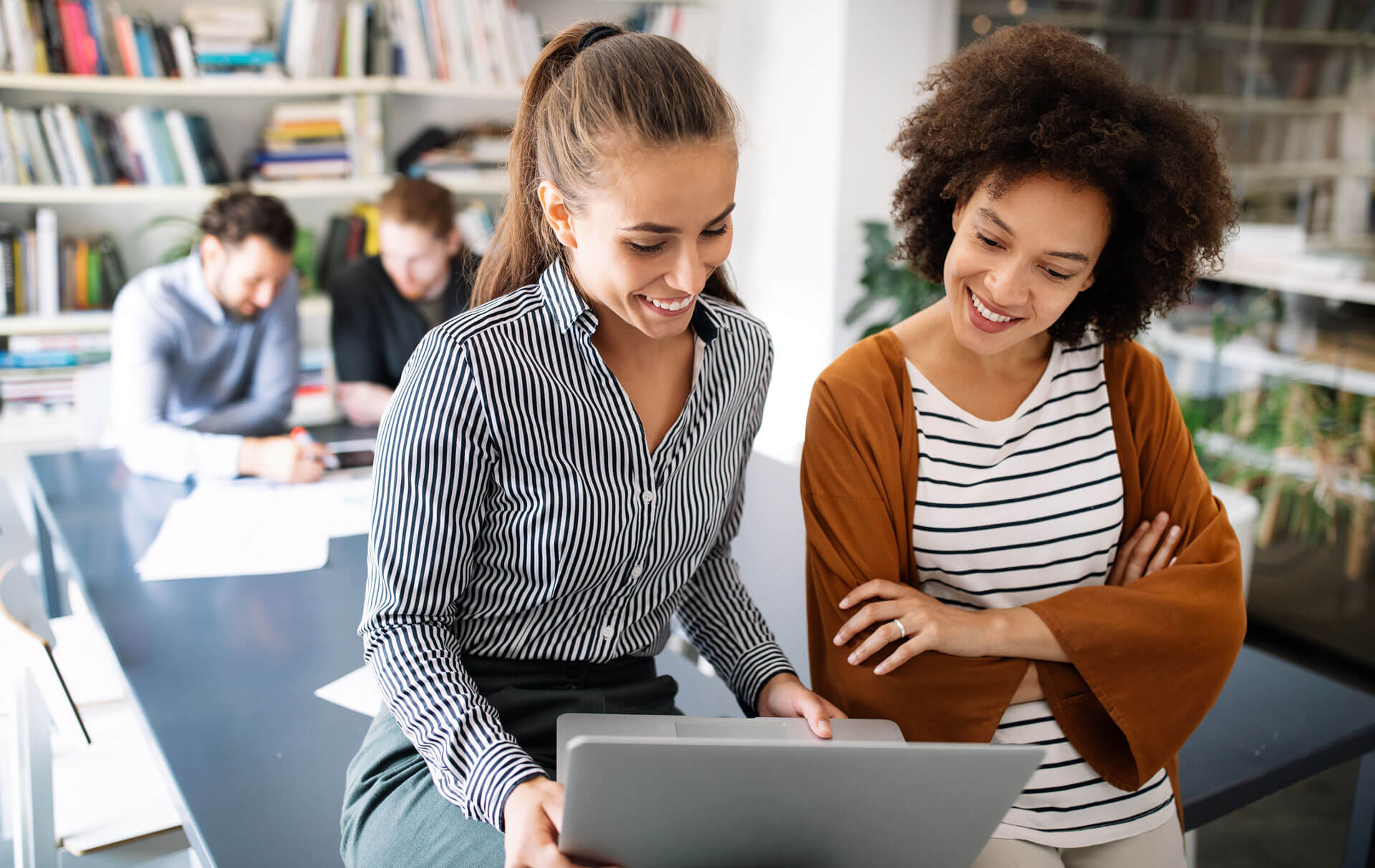 Two women are smiling and looking at a laptop screen in an office setting. One is wearing a striped shirt, and the other a white top with a brown cardigan. In the background, two people are sitting at a desk, focused on their work. Bookshelves line the walls.