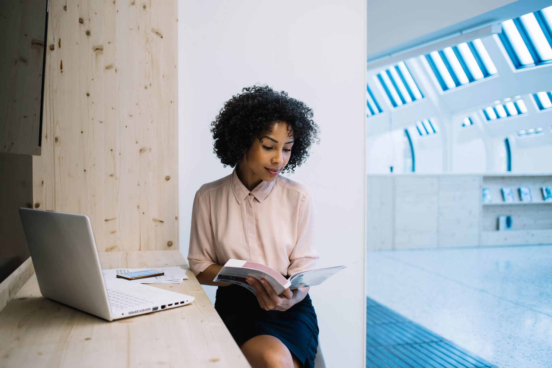 A woman with curly hair sits in a bright, modern space, reading a book. She's dressed in a light pink shirt and black skirt. A laptop and smartphone are on the wooden surface beside her, with large windows in the background.