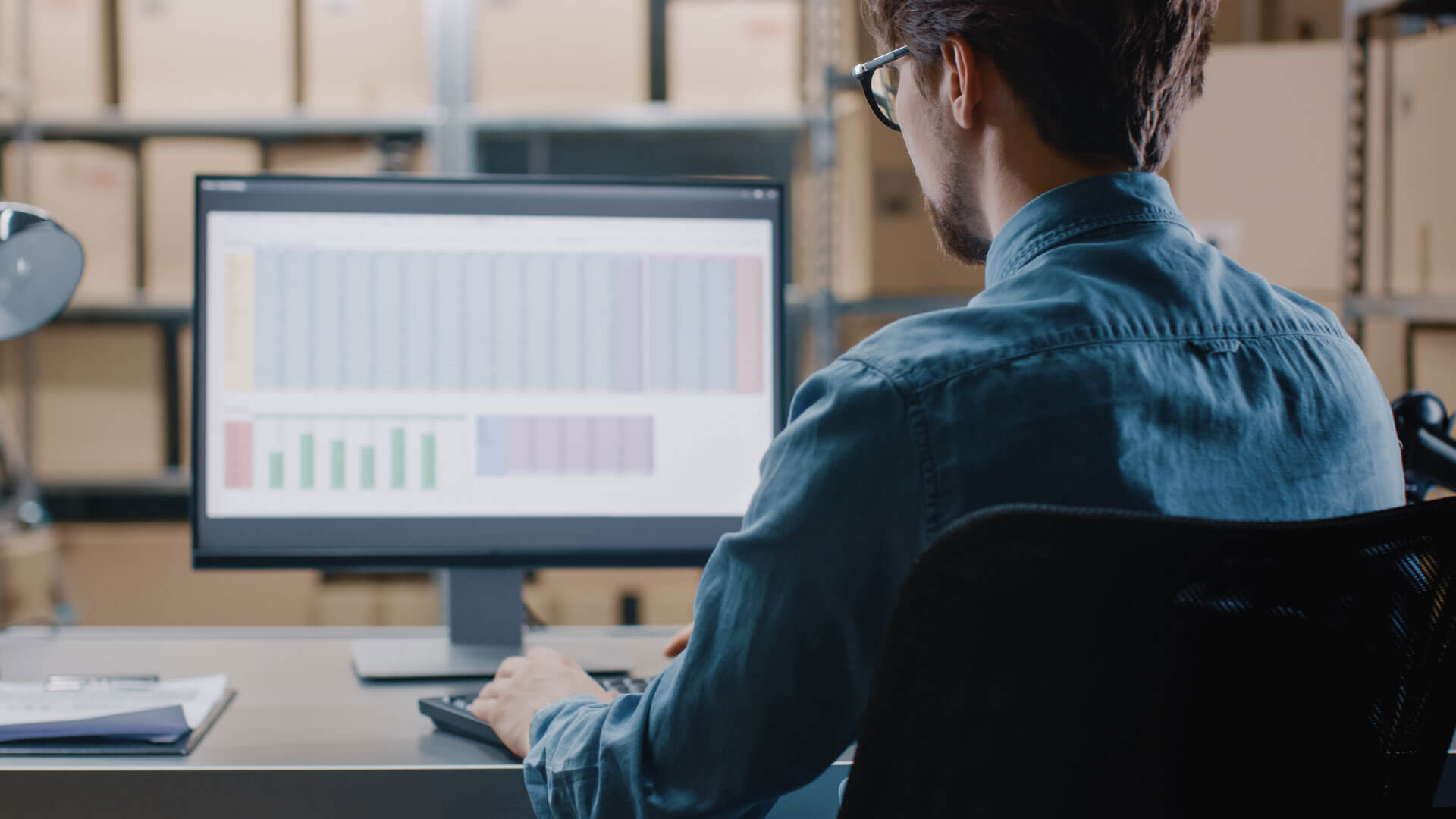 A person in a denim shirt sits at a desk, working on a computer with a spreadsheet displayed on the screen. Shelves with boxes are blurred in the background.