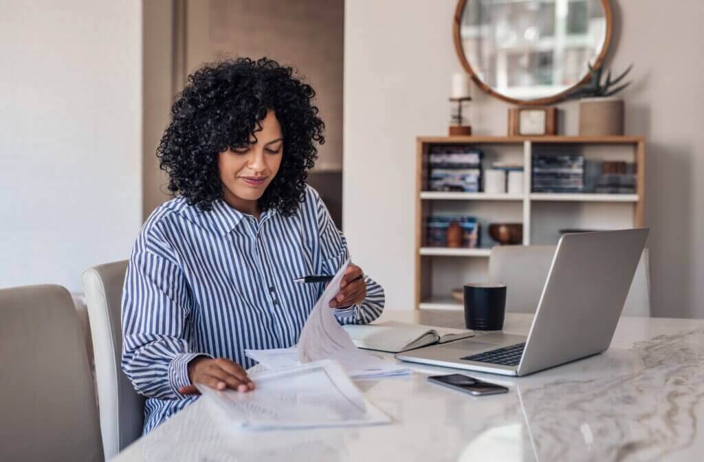 A woman with curly hair sits at a marble table, reviewing papers. She wears a striped shirt and holds a pen. A laptop, smartphone, and coffee mug are on the table. Shelves with books and decor are in the background.