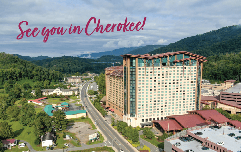 Aerial view of a large hotel surrounded by green hills and buildings, with a road leading through the area. The sky is partly cloudy. Text overlay reads, "See you in Cherokee!.