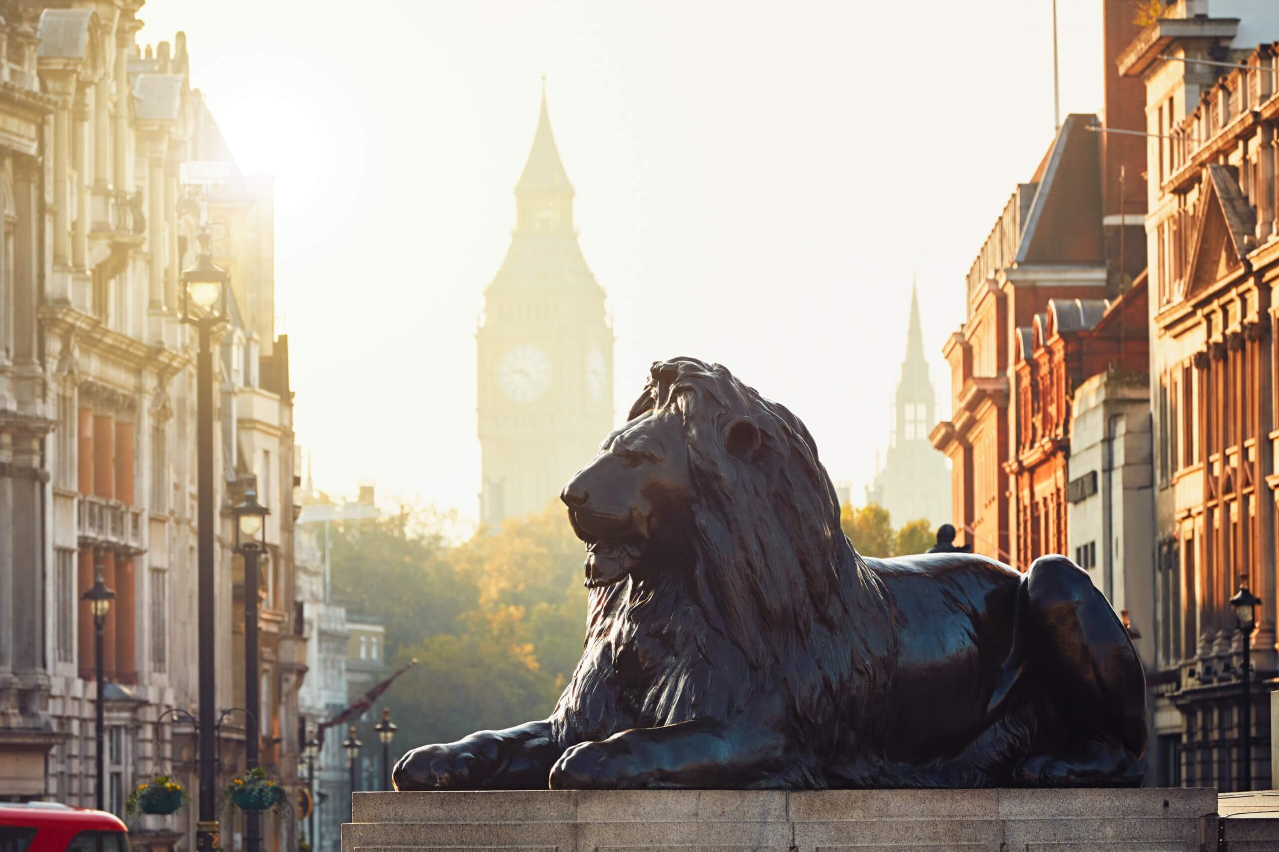 A bronze lion sculpture rests on a plinth in the foreground with the clock tower, commonly known as Big Ben, in the background. Sunlight creates a warm glow, illuminating the historic buildings lining the street.