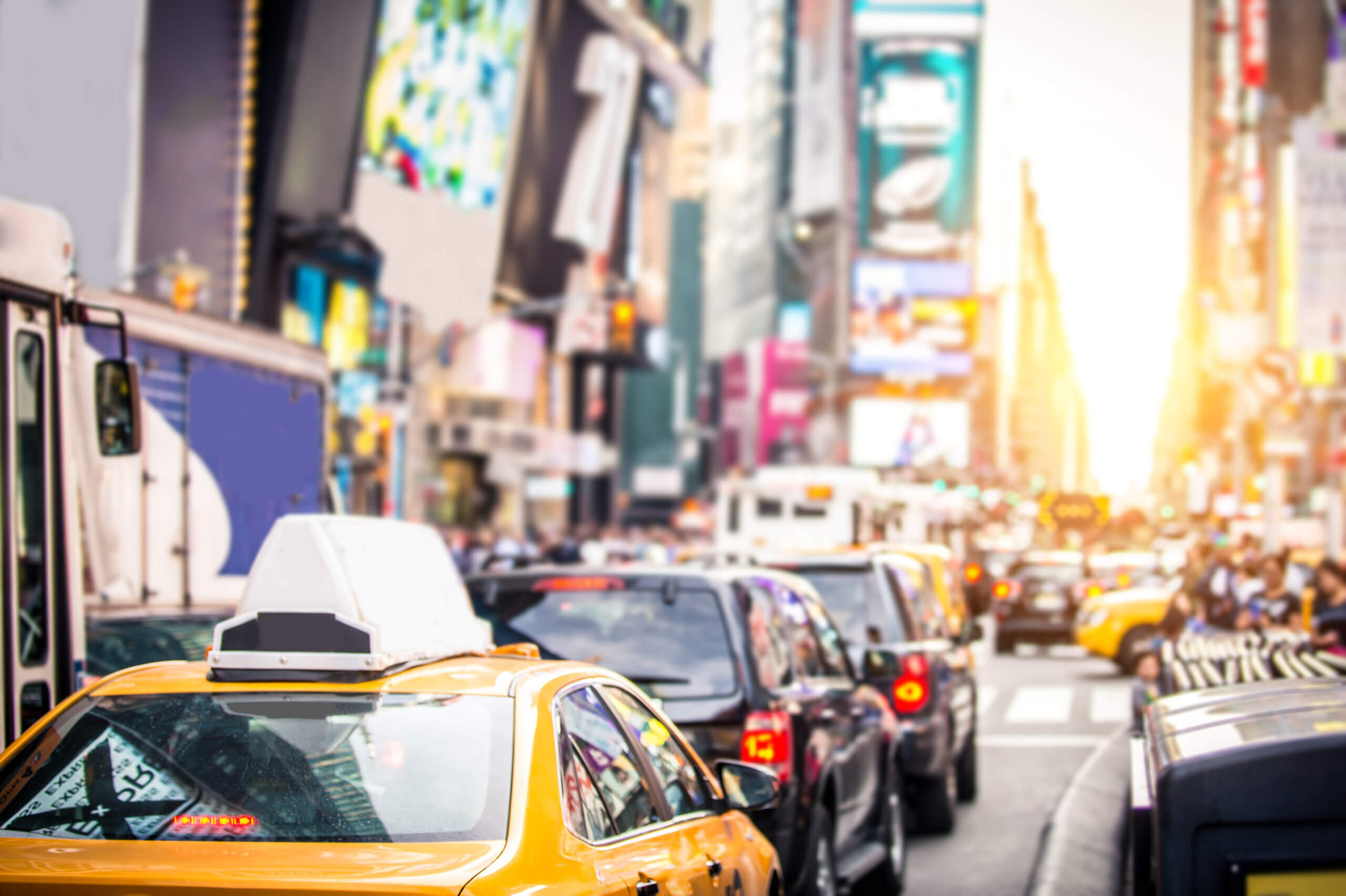 A bustling city street during sunset with yellow taxis and other vehicles in traffic. The background features bright, blurred billboards and tall buildings in the distance.