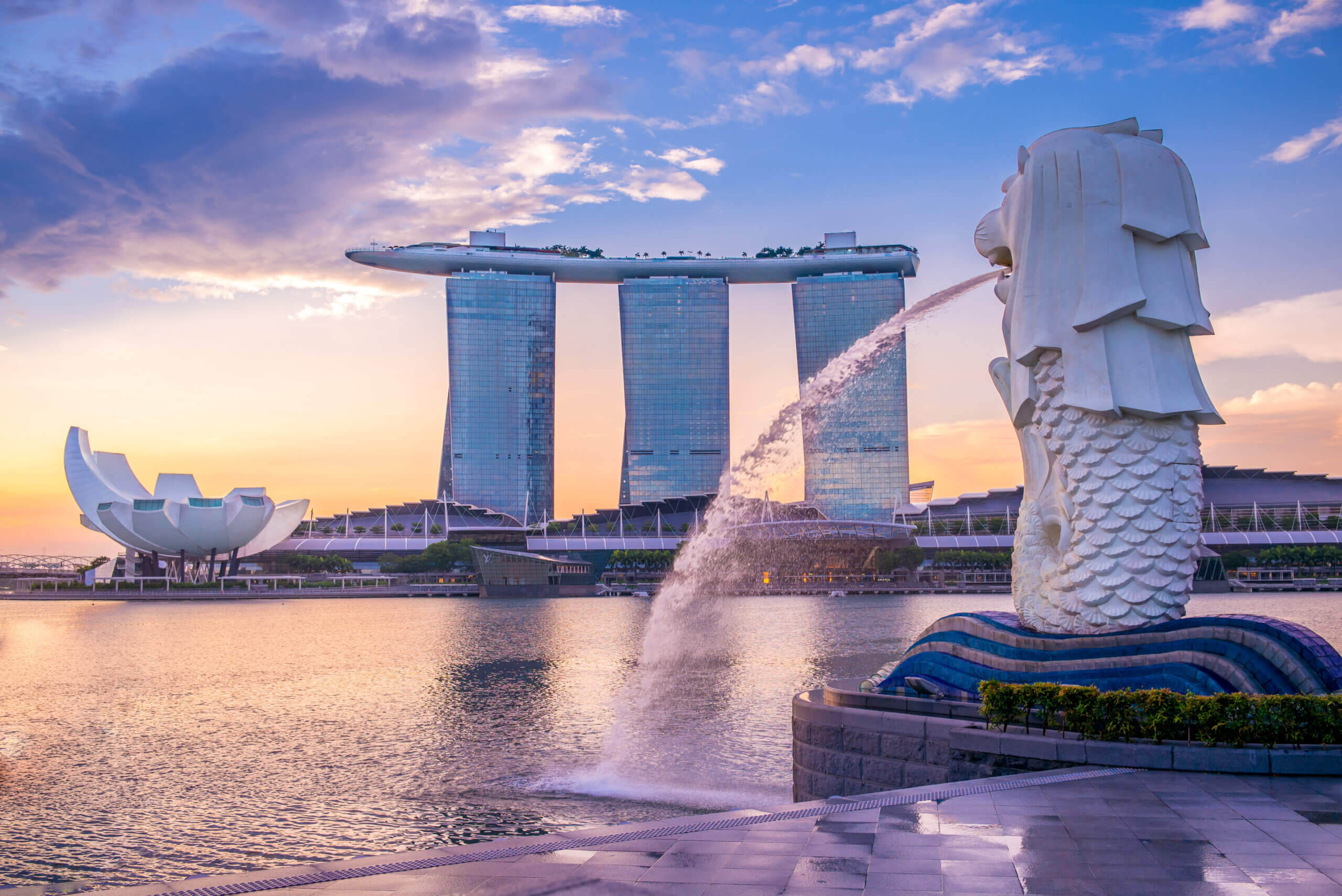 The image shows the Merlion statue in Singapore overlooking Marina Bay. In the background is the Marina Bay Sands hotel with its iconic rooftop, under a vibrant sky during sunrise or sunset.